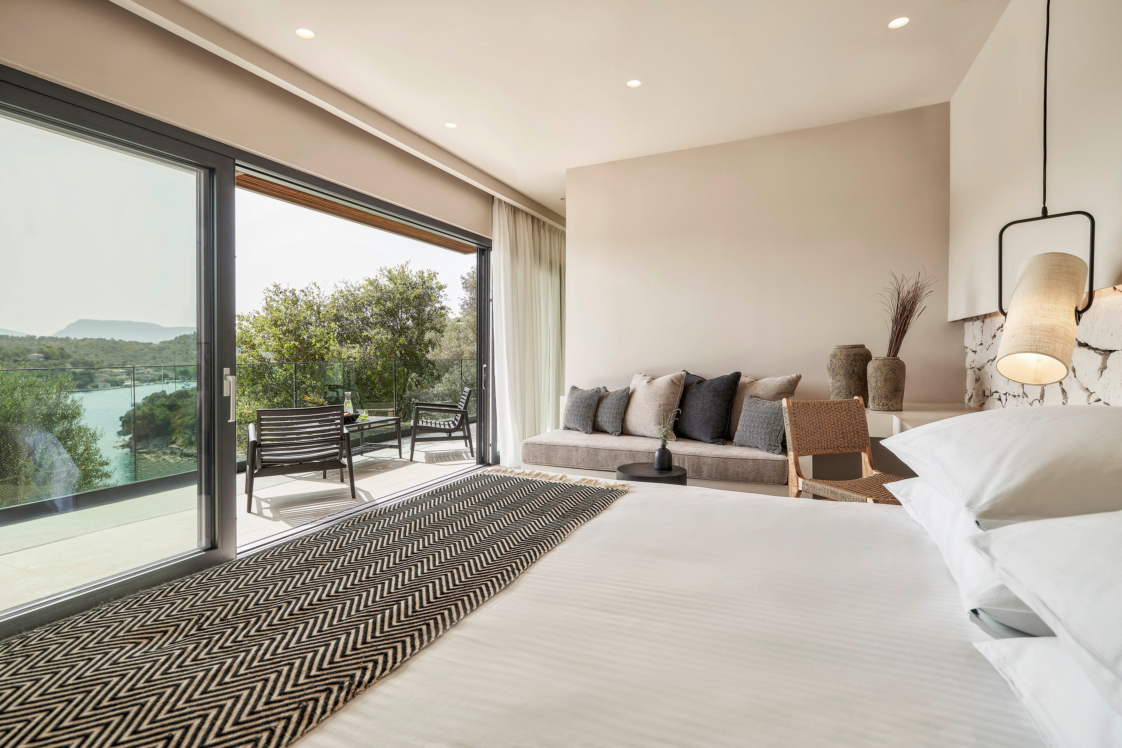 A contemporary bedroom featuring a neatly made bed, a patterned runner, and a cozy seating area. The room opens to a balcony with outdoor furniture and a scenic view of greenery and water. Neutral tones and soft lighting create a serene and inviting atmosphere.