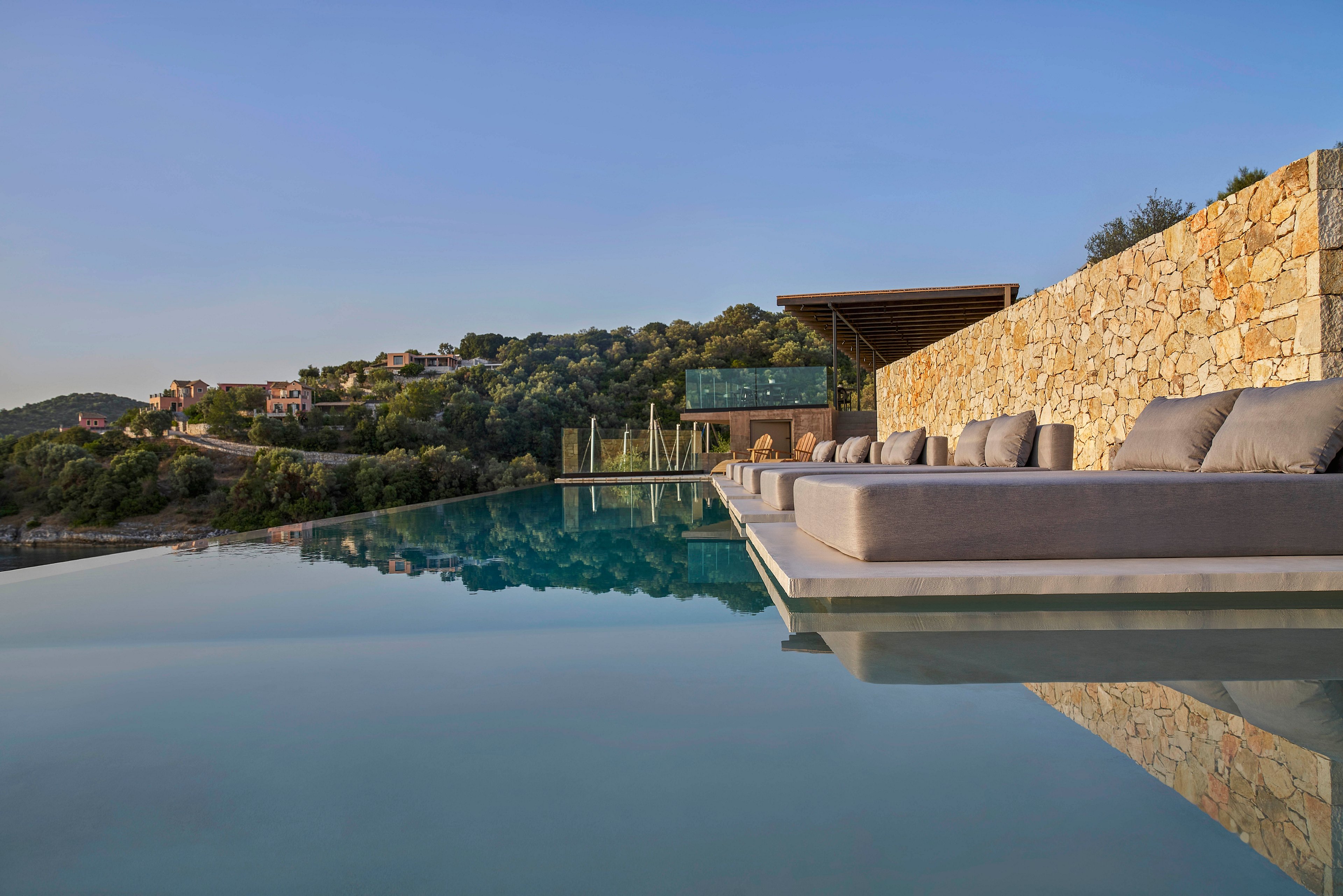A serene infinity pool surrounded by modern stone architecture overlooks a lush hillside. Comfortable seating is arranged along the poolside, creating a relaxing atmosphere. The setting is bathed in soft natural light, enhancing the tranquil mood.