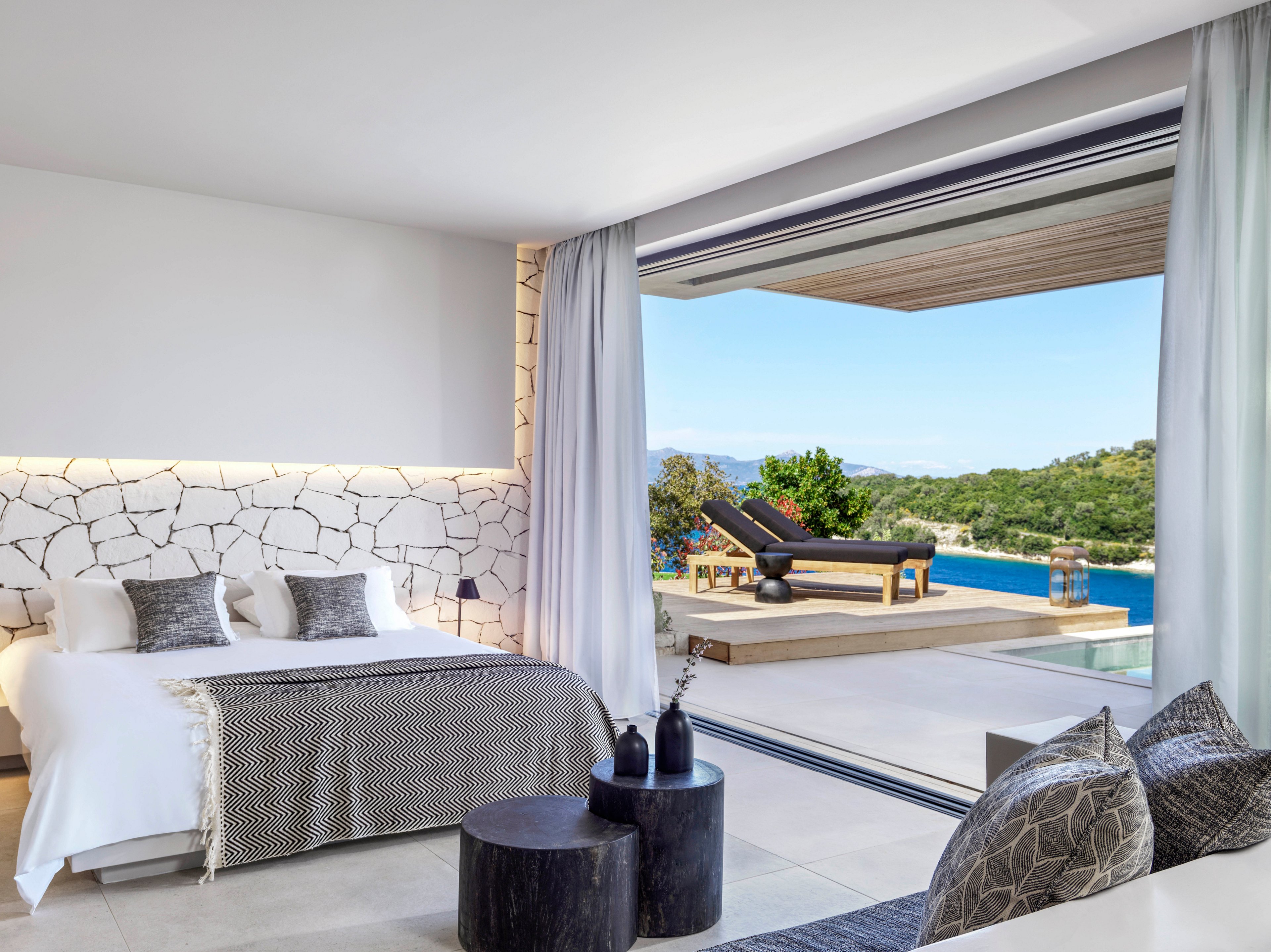 A modern, luxurious bedroom featuring a stone-accented wall and a neatly made bed with decorative pillows. The room opens to a spacious terrace with lounge chairs overlooking a serene ocean view and lush greenery. The neutral color palette and natural light create a tranquil and inviting atmosphere.