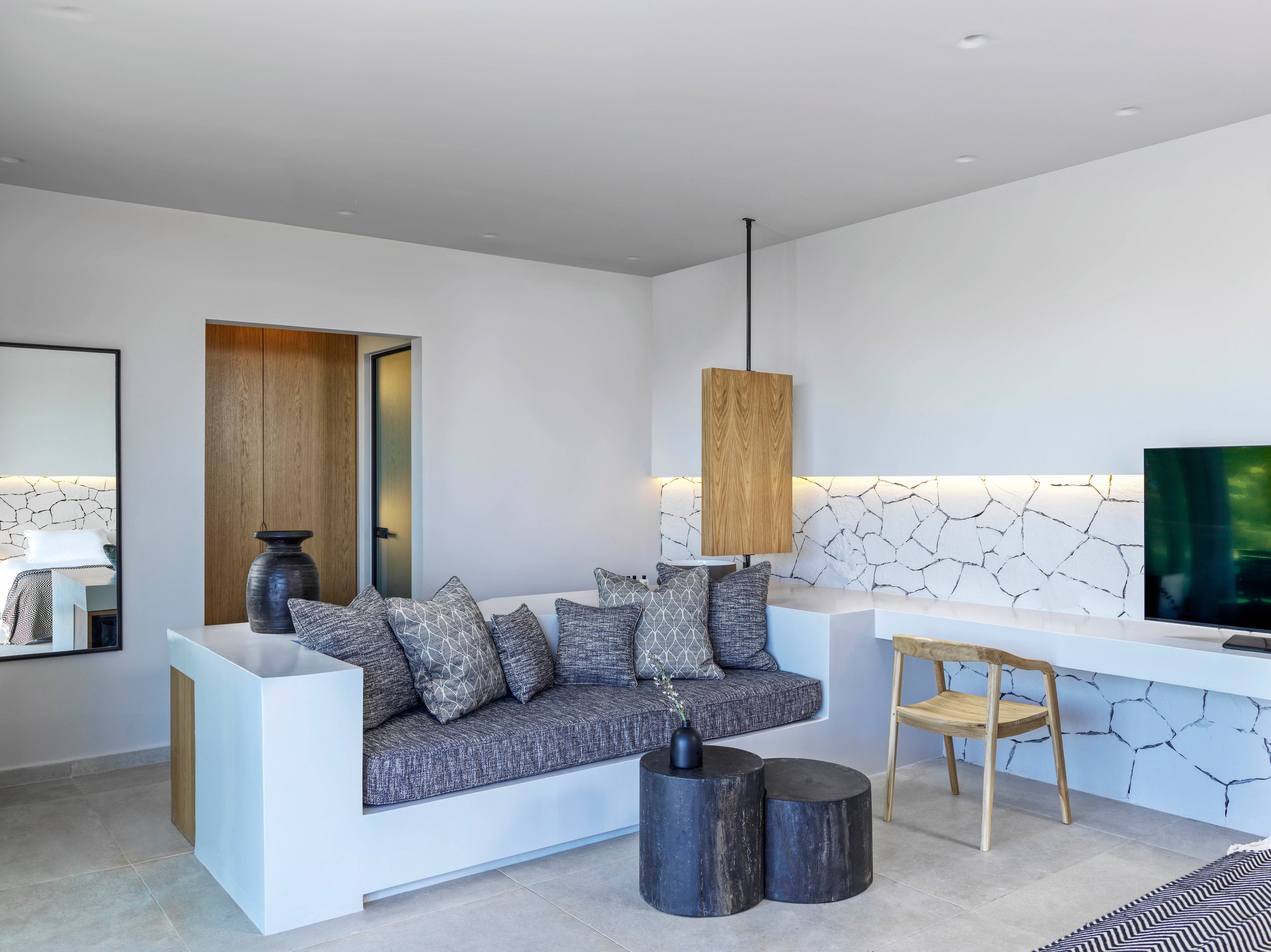 A contemporary living room featuring a minimalist design with a built-in sofa adorned with patterned cushions. The space includes a sleek wooden desk, a flat-screen TV, and decorative elements like vases and pendant lighting. The white walls and marble-patterned backsplash add a clean and sophisticated touch.