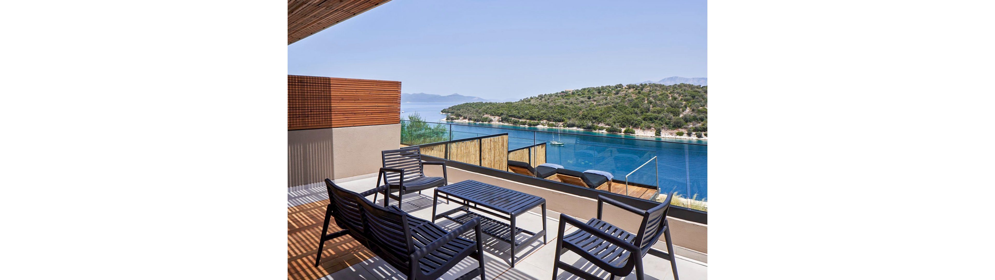 A contemporary terrace features black metal outdoor chairs and a matching table facing a serene blue bay. Glass railings and wooden privacy panels frame the view of the water and green hillside across the inlet. Two sun loungers are positioned toward the sea, suggesting a relaxing vacation setting. The clear sky and calm water create a peaceful, minimalist atmosphere.