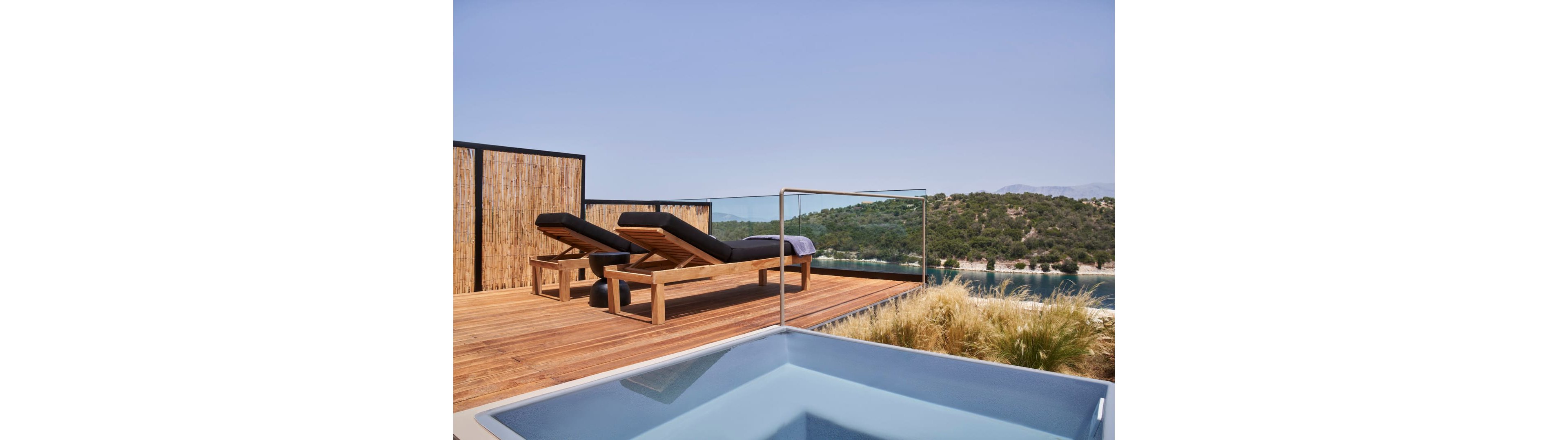 A contemporary rooftop terrace features two wooden sun loungers with dark cushions facing a scenic landscape. The deck is made of warm-toned wood and bordered by a natural reed privacy screen. In the foreground, a sleek square pool or hot tub with clear water adds a luxurious touch. Beyond the glass railing, green hills and a calm body of water create a tranquil, resort-like atmosphere under a clear sky.