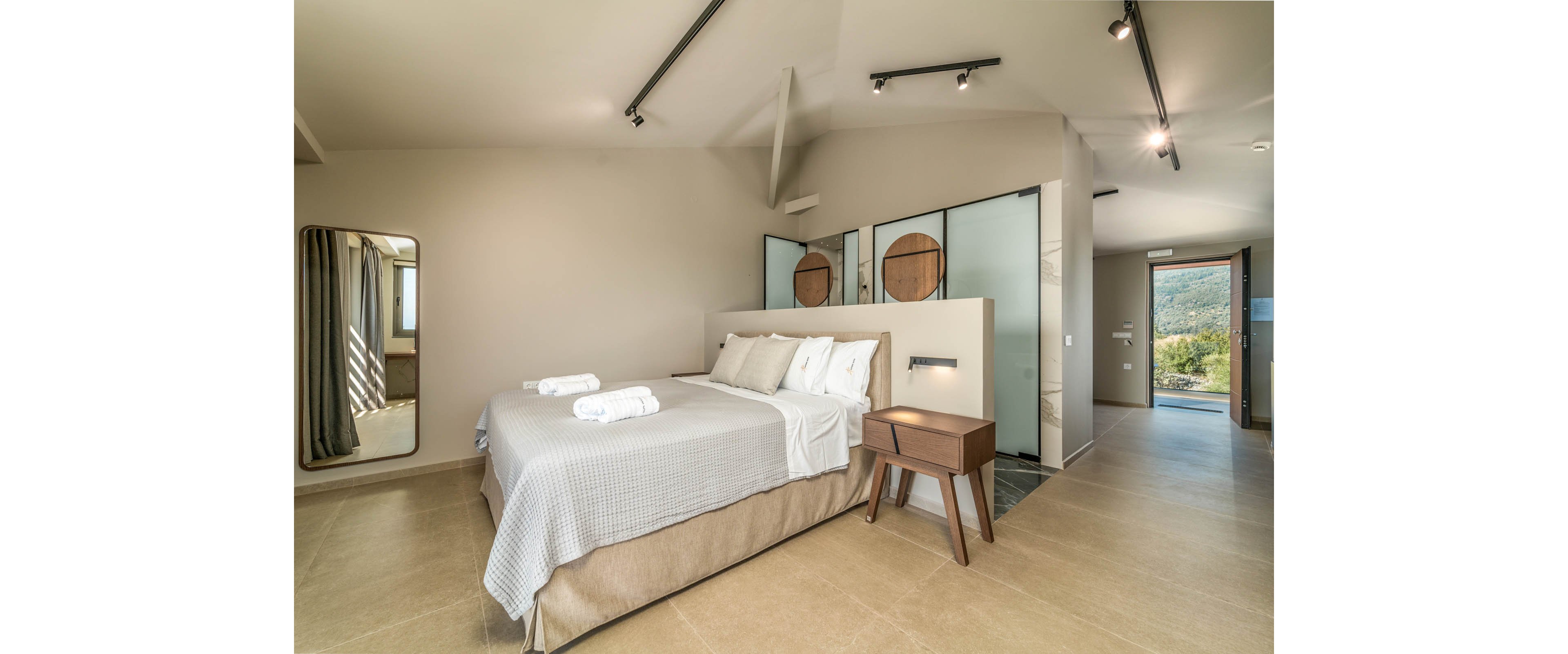 A contemporary bedroom features a neatly made double bed with folded white towels placed on top. Neutral tones dominate the space, with beige flooring, light walls, and simple bedding creating a calm atmosphere. A wooden bedside table, large wall mirror, and glass partition add modern design elements. Natural light enters from the open doorway, revealing greenery and hills outside for a serene, inviting setting.