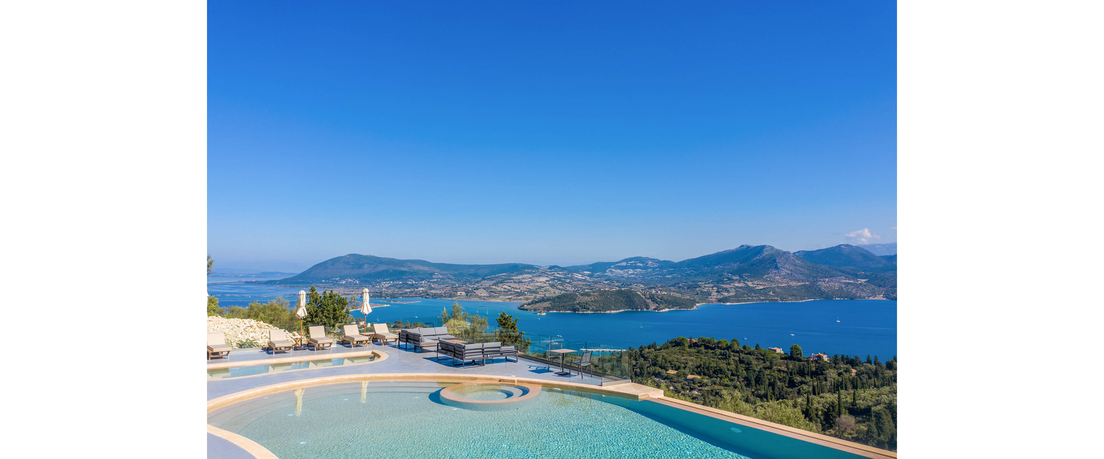 A luxurious infinity pool curves gracefully in the foreground, reflecting the clear blue sky. Sun loungers and outdoor seating are arranged around the pool, suggesting a relaxing resort or private villa setting. Beyond the terrace, a sweeping view of a deep blue bay stretches toward distant mountains and rolling hills. The bright, cloudless day and vivid colors create a serene and inviting vacation atmosphere.