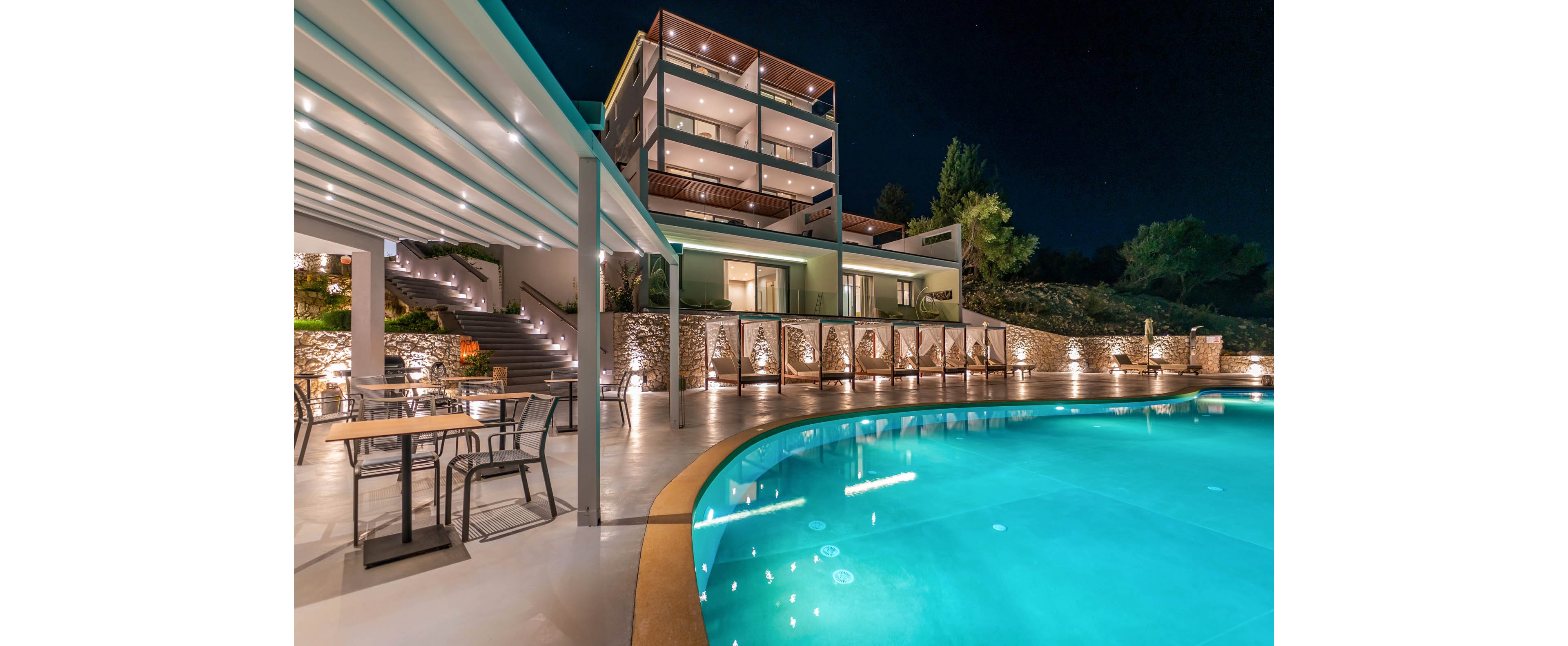 A contemporary multi-story hotel overlooks a curved swimming pool glowing with turquoise light at night. The scene includes an open-air terrace with tables and chairs under a sleek pergola. Warm accent lighting along stone walls and stairways creates an inviting atmosphere. Trees and landscaping frame the property against a dark, clear sky.