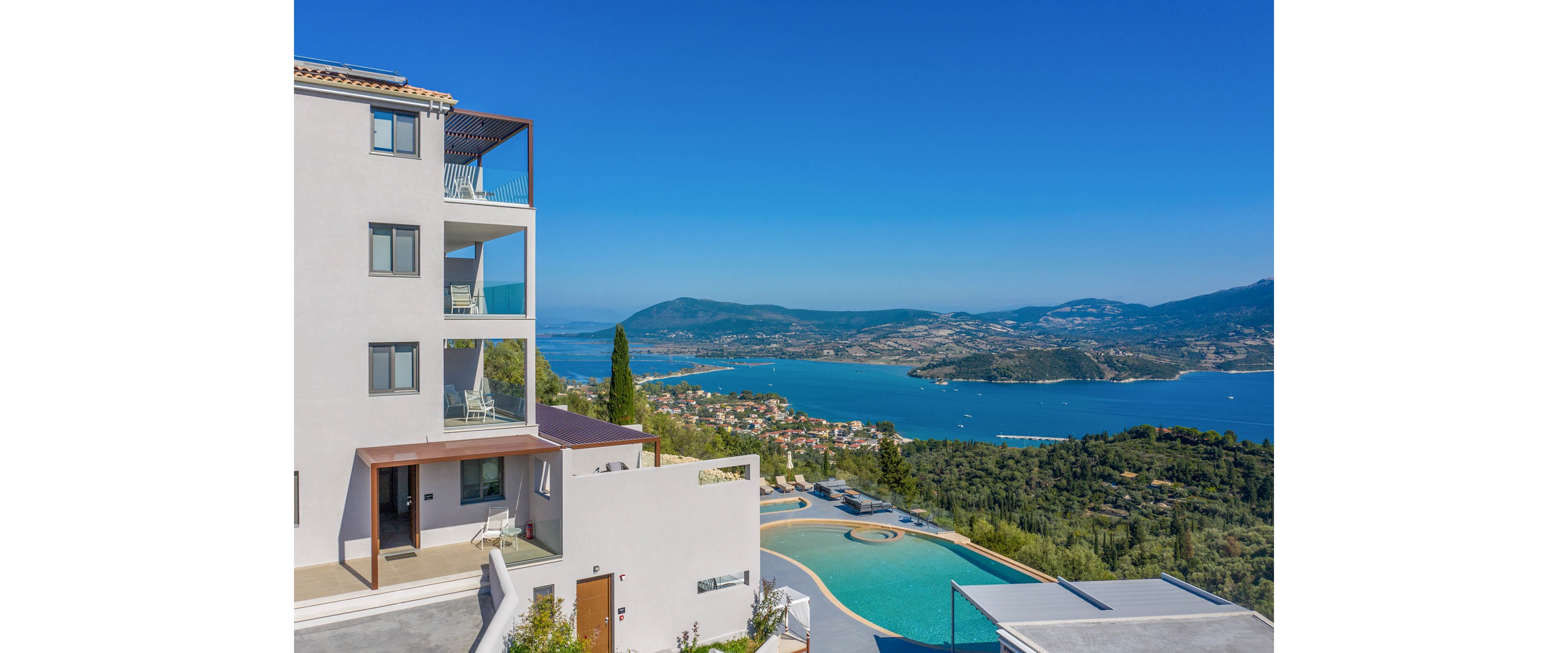 A contemporary multi-story villa stands on a hillside with expansive glass balconies and terraces. Below, an infinity-style swimming pool curves along the edge of the property, framed by greenery and stone. The scene overlooks a wide coastal bay with blue water, distant mountains, and scattered buildings along the shoreline. Clear skies and bright sunlight create a serene, luxurious vacation atmosphere.