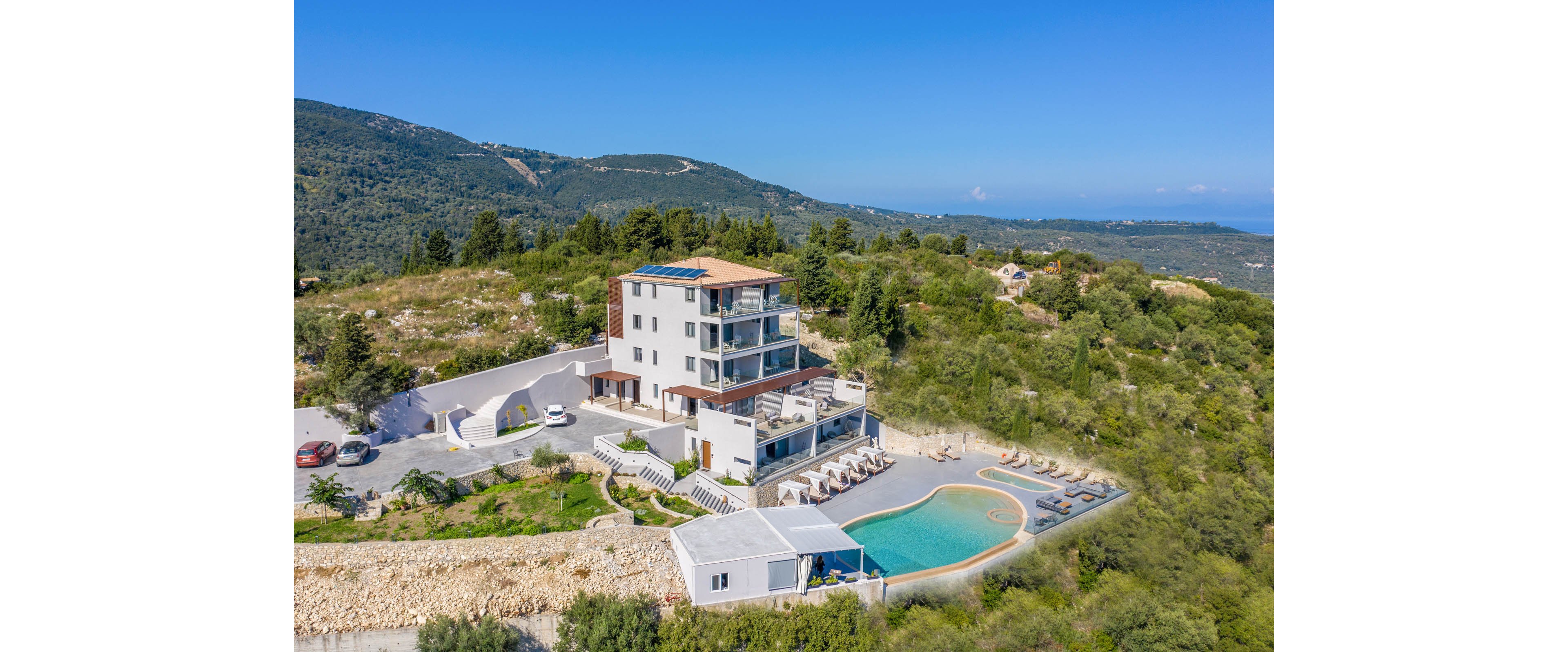 A modern multi-story hotel stands on a hilltop surrounded by lush green landscape and distant mountains. The property features an outdoor infinity-style swimming pool with sun loungers neatly arranged along the poolside terrace. Several parked cars and a paved driveway indicate guest access and arrival. Clear blue skies and expansive natural scenery create a serene vacation atmosphere.