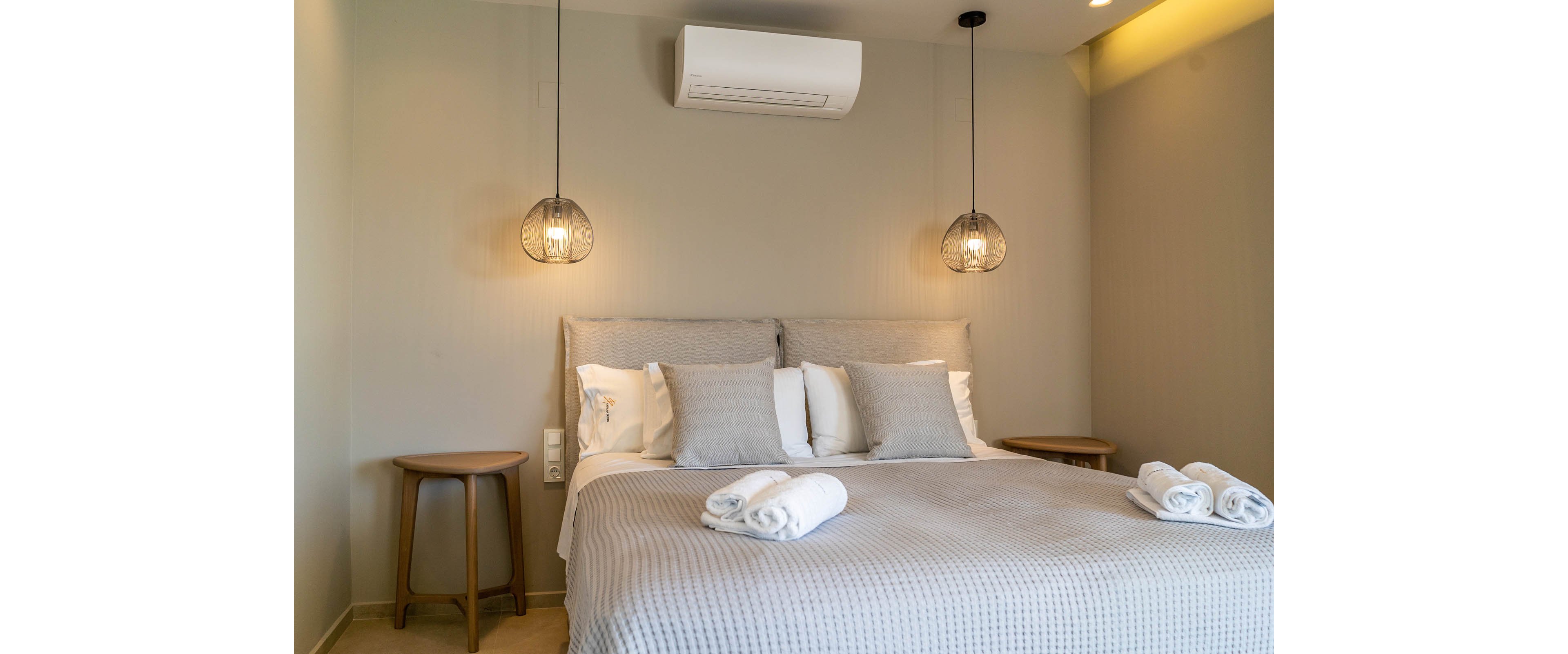 A neatly made double bed with neutral bedding is centered in a modern bedroom. Two pendant lights hang above matching wooden bedside tables on either side of the bed. Rolled white towels are placed at the foot of the bed, suggesting a hotel or guest room setting. Soft, warm lighting and minimal decor create a calm, contemporary atmosphere.