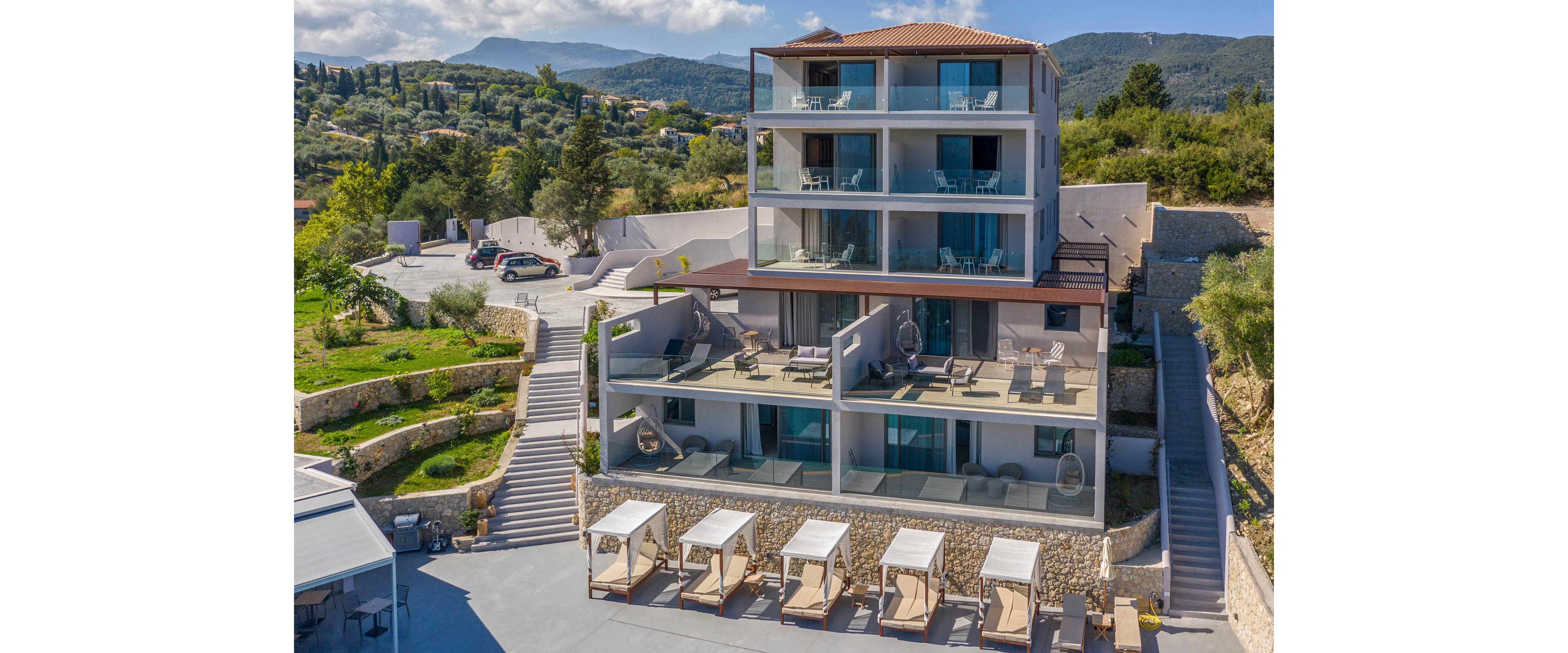 A contemporary multi-story resort building stands on a hillside with expansive terraces and balconies overlooking the surrounding landscape. Sun loungers and cabanas are arranged neatly in the foreground near a paved area, suggesting a relaxing vacation setting. Stone retaining walls, stairways, and landscaped greenery frame the property, leading up to the main structure. Rolling hills and distant mountains complete the scenic backdrop under a clear sky.