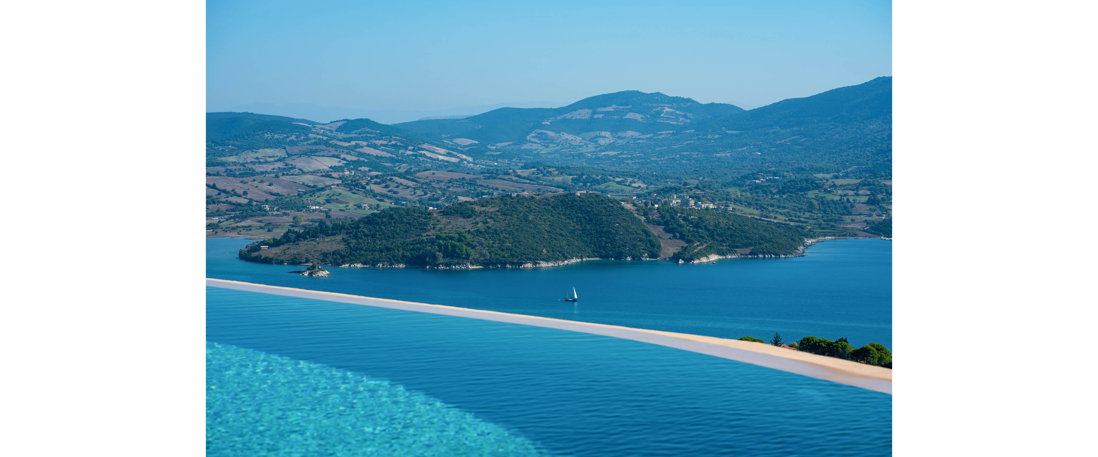 A serene infinity pool stretches across the foreground, blending seamlessly into a calm blue sea. In the distance, a small sailboat glides across the water near a lush green island. Rolling hills and mountains form a layered backdrop under a clear blue sky. The scene evokes relaxation and luxury with vibrant turquoise and deep blue tones.