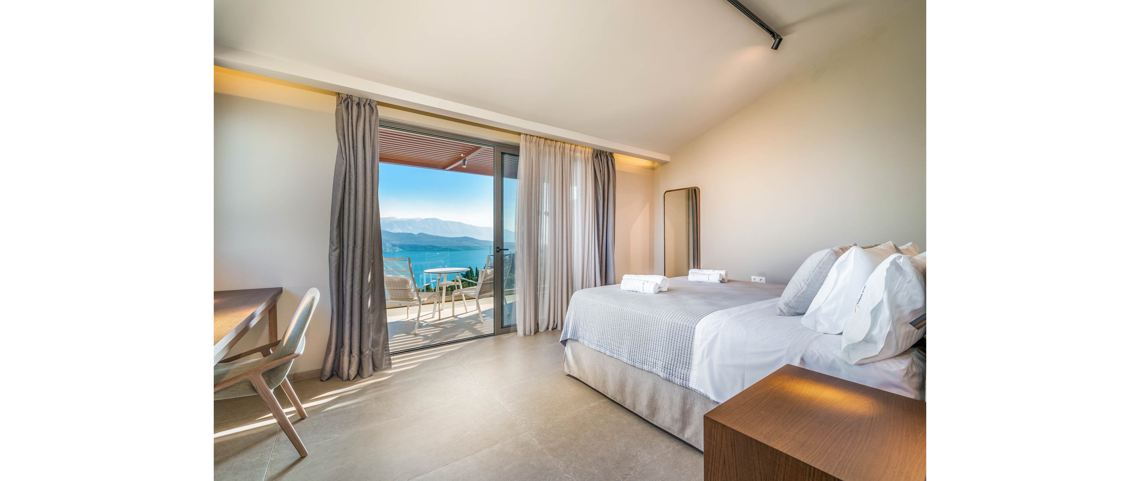Modern hotel bedroom with a neatly made double bed faces a large sliding glass door. The room opens onto a balcony with chairs overlooking a blue sea and distant mountains. Neutral tones, soft curtains, and minimal furniture create a clean, contemporary atmosphere. Sunlight fills the space, emphasizing the relaxing vacation setting.