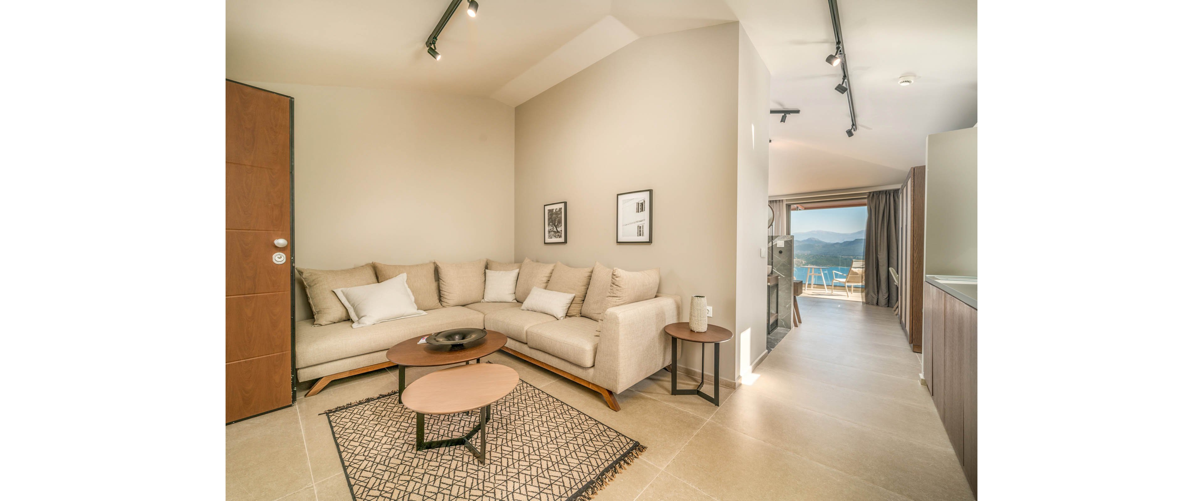 Contemporary living room interior featuring a beige sectional sofa and minimalist decor. The space includes nested round coffee tables on a patterned rug and framed artwork on the wall. Track lighting on the ceiling illuminates the room, leading toward a bright balcony with mountain and water views. Neutral tones and clean lines create a calm, modern atmosphere.
