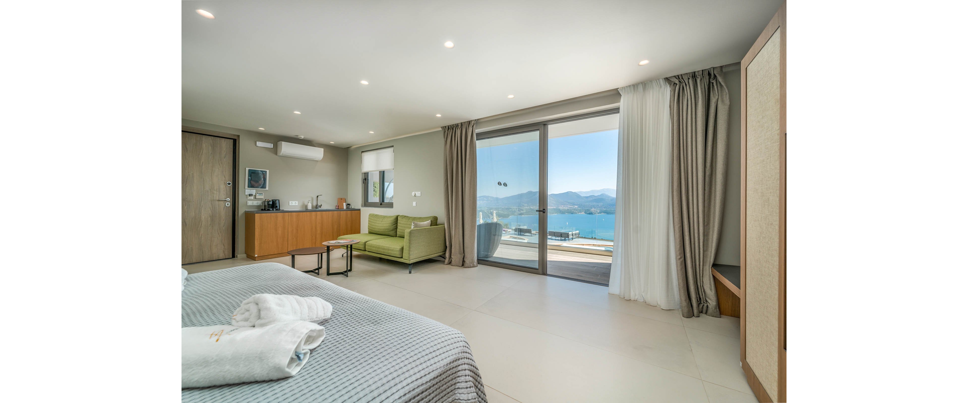 Spacious contemporary hotel room features a neatly made bed in the foreground and a sitting area with a green sofa. A kitchenette with wooden cabinetry and a wall-mounted air conditioner lines one side of the room. Large sliding glass doors open to a balcony overlooking a bright blue sea and distant mountains under clear skies. Neutral tones, minimal decor, and ample natural light create a calm, relaxing atmosphere.
