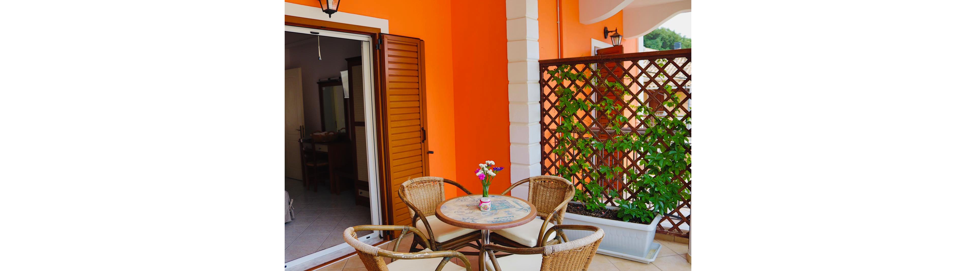 A small balcony patio features a round table with four wicker chairs arranged around it. The bright orange exterior wall and white column create a vibrant backdrop next to an open wooden shutter door leading indoors. A lattice screen supports green climbing plants in a long planter, adding natural texture and privacy. A small vase with colorful flowers sits on the tabletop, enhancing the inviting, relaxed atmosphere of the outdoor space.
