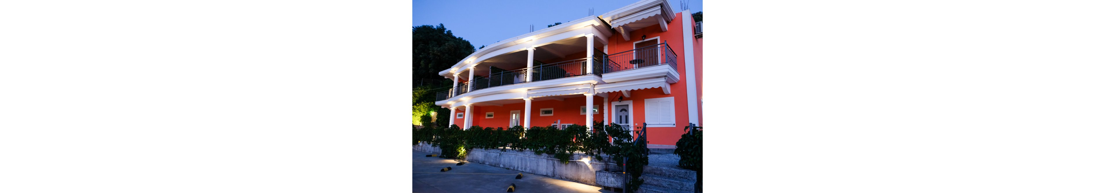 A two-story coral-colored villa is shown in the evening, with bright exterior lights highlighting its balconies and white columns. The house features large terraces with metal railings and an awning-style roofline. Green shrubs line the front of the property above a stone retaining wall, and a paved driveway or courtyard extends across the foreground. The sky is transitioning to twilight, giving the scene a calm, inviting atmosphere.
