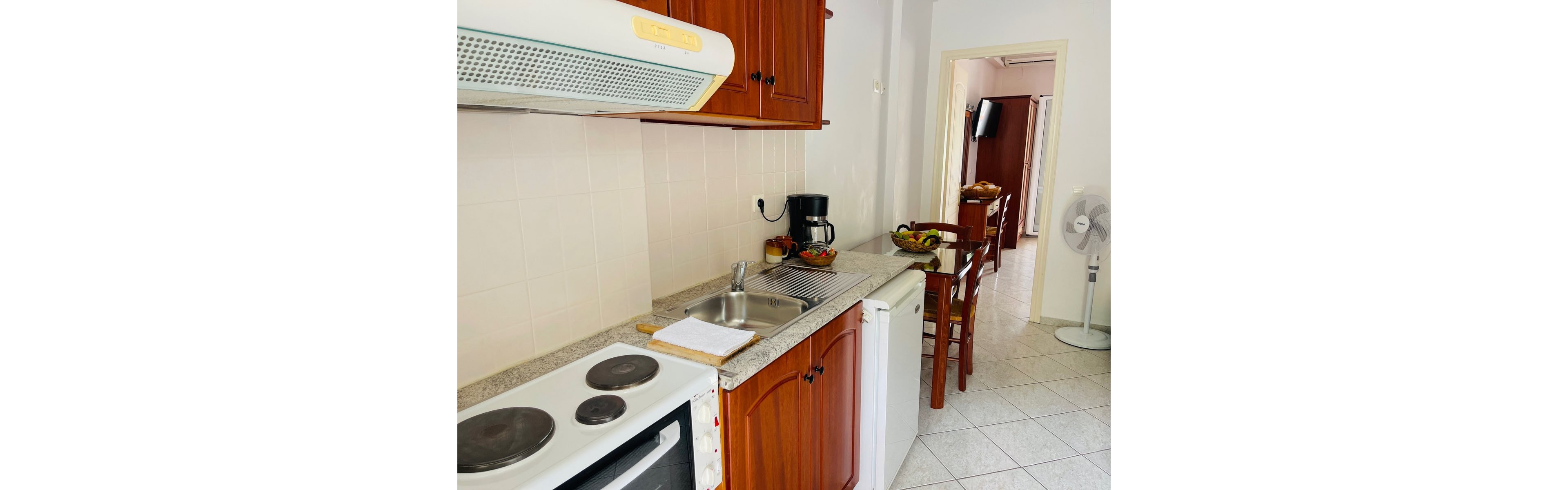 A small apartment kitchen features a white electric stove, stainless steel sink, and wooden cabinets along one wall. A coffee maker and fruit bowl sit on the countertop, adding color to the neutral tiled space. In the background, a wooden dining table with chairs is visible, along with a standing fan and a wall-mounted television in the adjoining room. The overall scene suggests a clean, functional vacation rental or studio apartment interior.