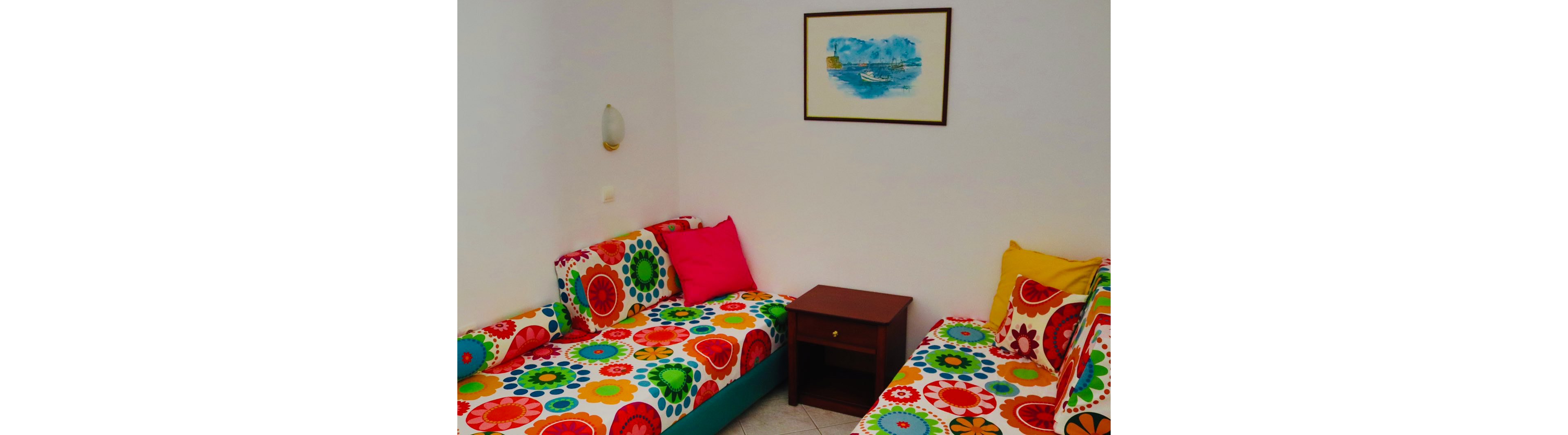 The image shows a small room with two twin sofas covered in bright, multicolored floral-patterned fabric. A wooden side table with a drawer stands between the sofas, adding a functional element to the seating area. The walls are plain white, with a wall-mounted light fixture and a framed painting of boats on blue water providing simple decoration. The overall scene feels cheerful and inviting, with bold colors contrasting against the neutral background.