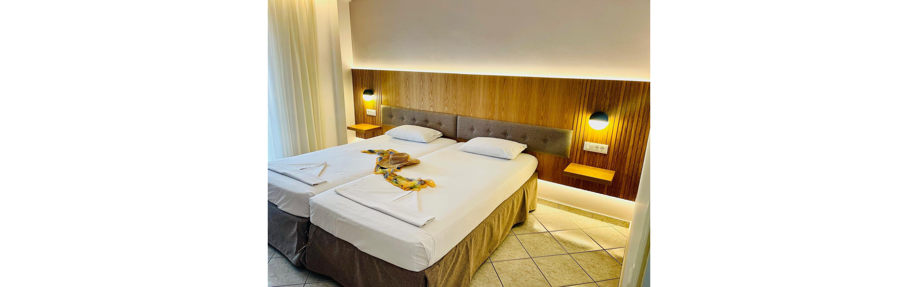 A bright, modern hotel room features two neatly made twin beds with white linens and beige bed skirts. A wooden headboard with upholstered panels runs along the wall, accented by two round wall lights casting a warm glow. Decorative towels and fabric are arranged on the beds, adding a welcoming touch. The room has tiled flooring and sheer curtains that suggest a window or balcony letting in natural light.