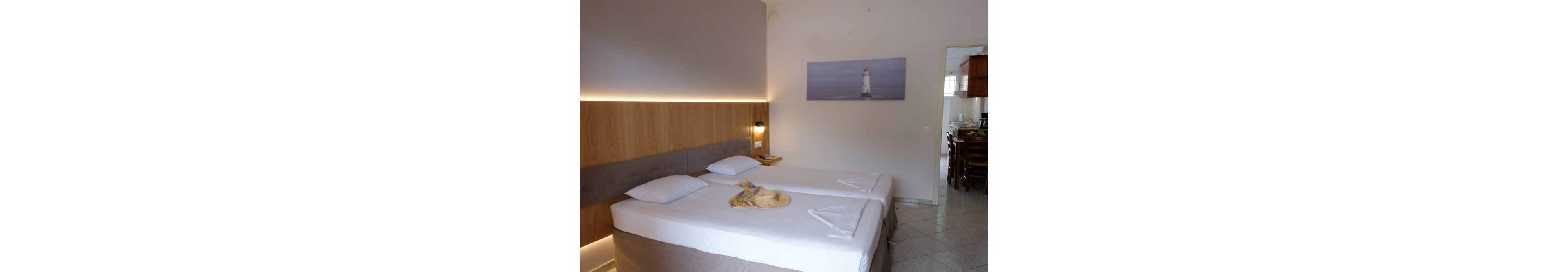 A modern hotel room features two neatly made twin beds with white linens and simple folded towel arrangements. A straw hat and colorful scarf rest on one of the beds, adding a casual vacation touch. The room has a wooden headboard with soft accent lighting and a wall-mounted bedside lamp. In the background, a doorway opens to a small dining or kitchen area, and a lighthouse artwork decorates the wall.