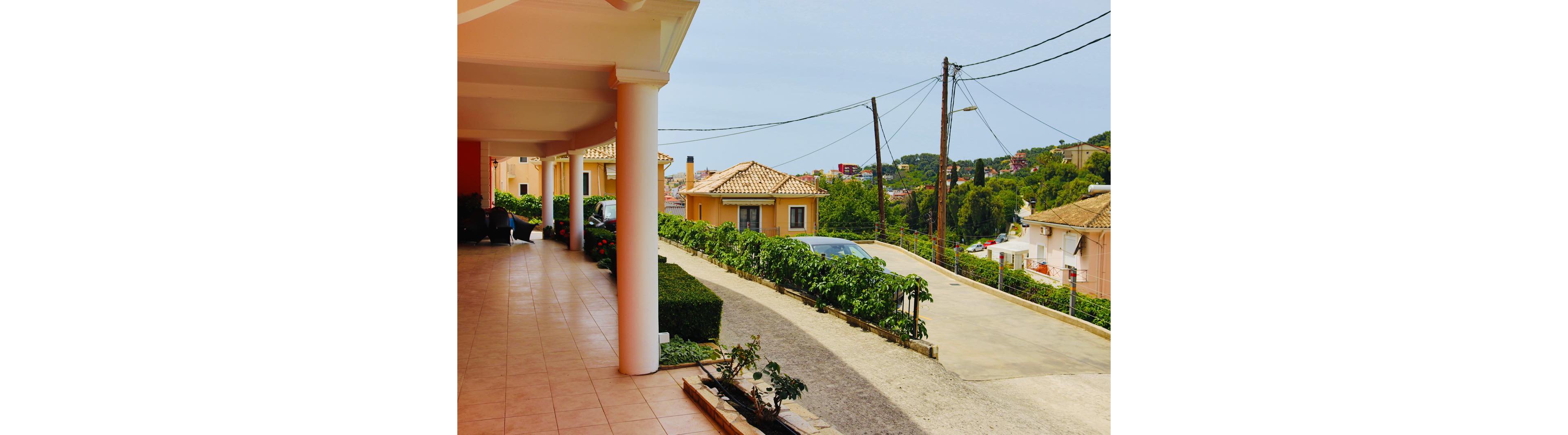 A bright tiled terrace with white columns overlooks a quiet residential street. Peach-colored houses with tiled roofs line the road, surrounded by greenery and shrubs. A parked car is visible near the center, and utility poles with overhead wires run along the street. The scene suggests a warm, peaceful neighborhood on a clear day.