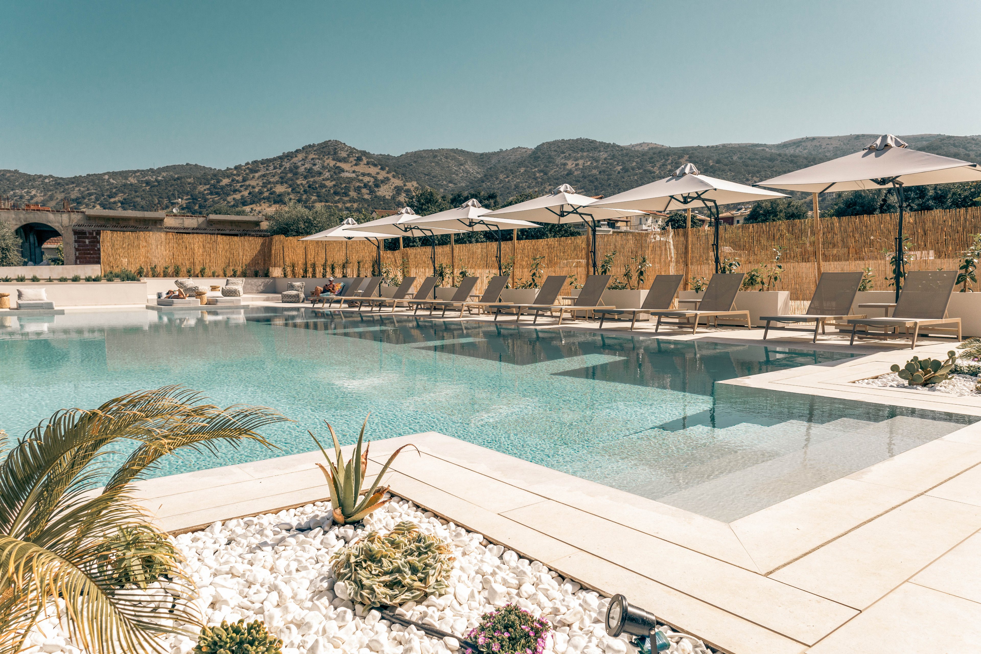 A serene outdoor swimming pool is surrounded by neatly arranged sun loungers and umbrellas at a luxury resort. The setting features light stone decking, decorative white gravel, and desert-style plants near the water’s edge. Mountains rise in the background under a clear blue sky, creating a tranquil vacation atmosphere. The overall scene feels calm, sunny, and inviting for relaxation.