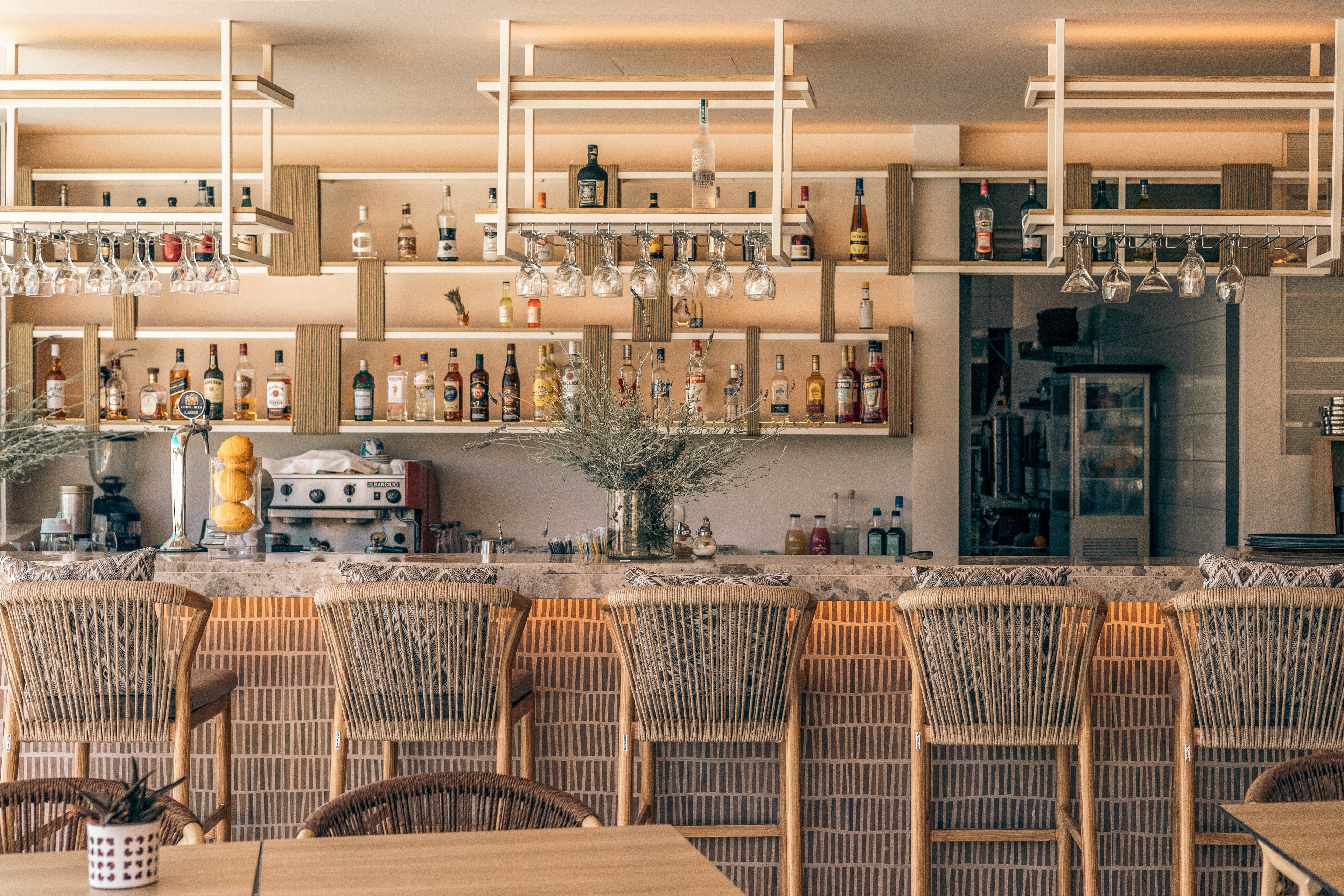 A contemporary bar interior features a long counter lined with wicker bar stools. Shelves behind the bar display an assortment of liquor bottles and hanging glassware in an organized arrangement. Warm ambient lighting and neutral tones create a cozy, inviting atmosphere. Decorative plants and neatly arranged equipment add to the clean, modern aesthetic of the space.
