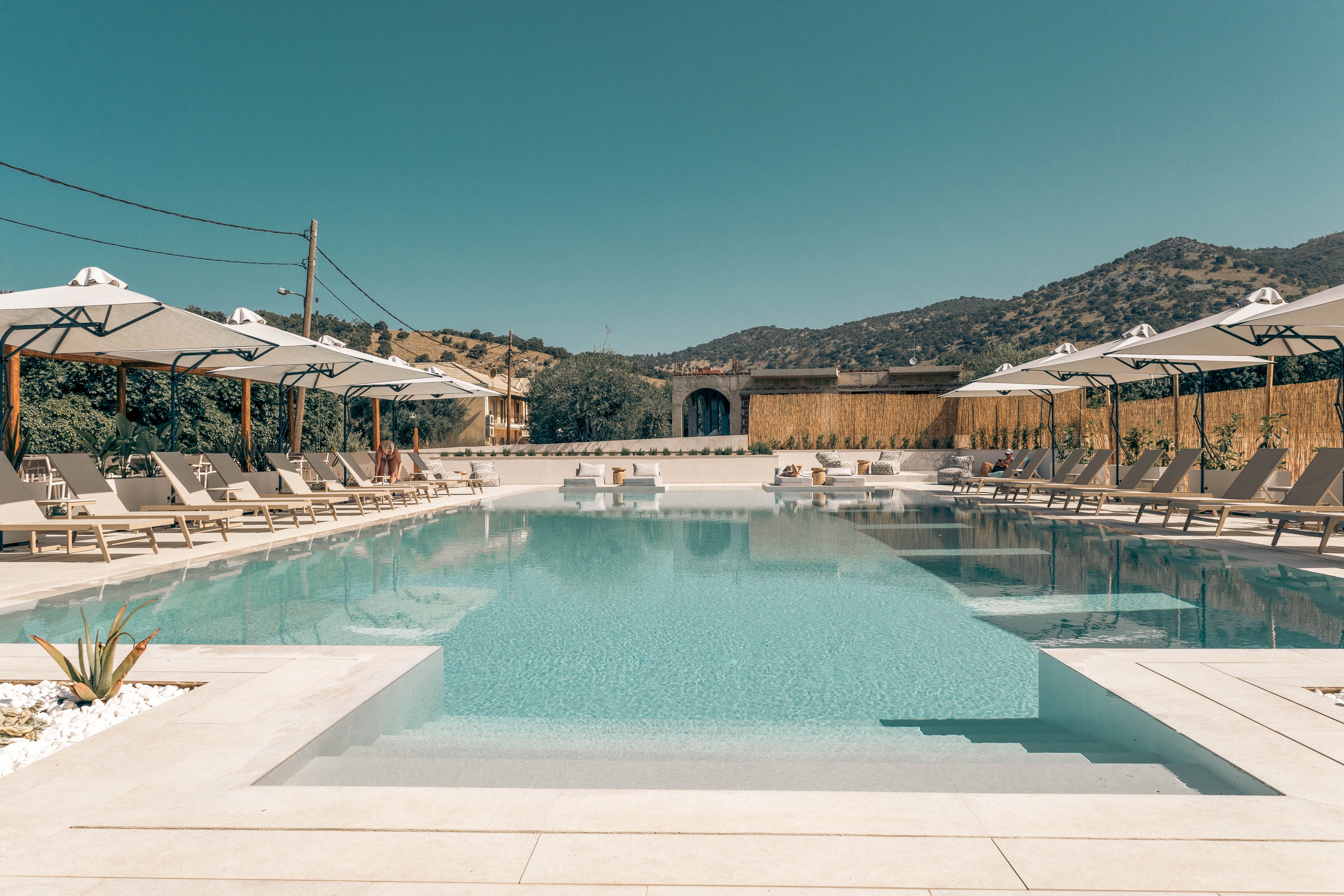 A serene outdoor swimming pool is centered between rows of sun loungers and large white umbrellas at a stylish resort. The clear blue water reflects the cloudless sky, creating a calm and inviting atmosphere. Minimalist stone decking surrounds the pool, with a few plants adding natural accents. Hills and buildings in the background suggest a scenic, secluded vacation setting.