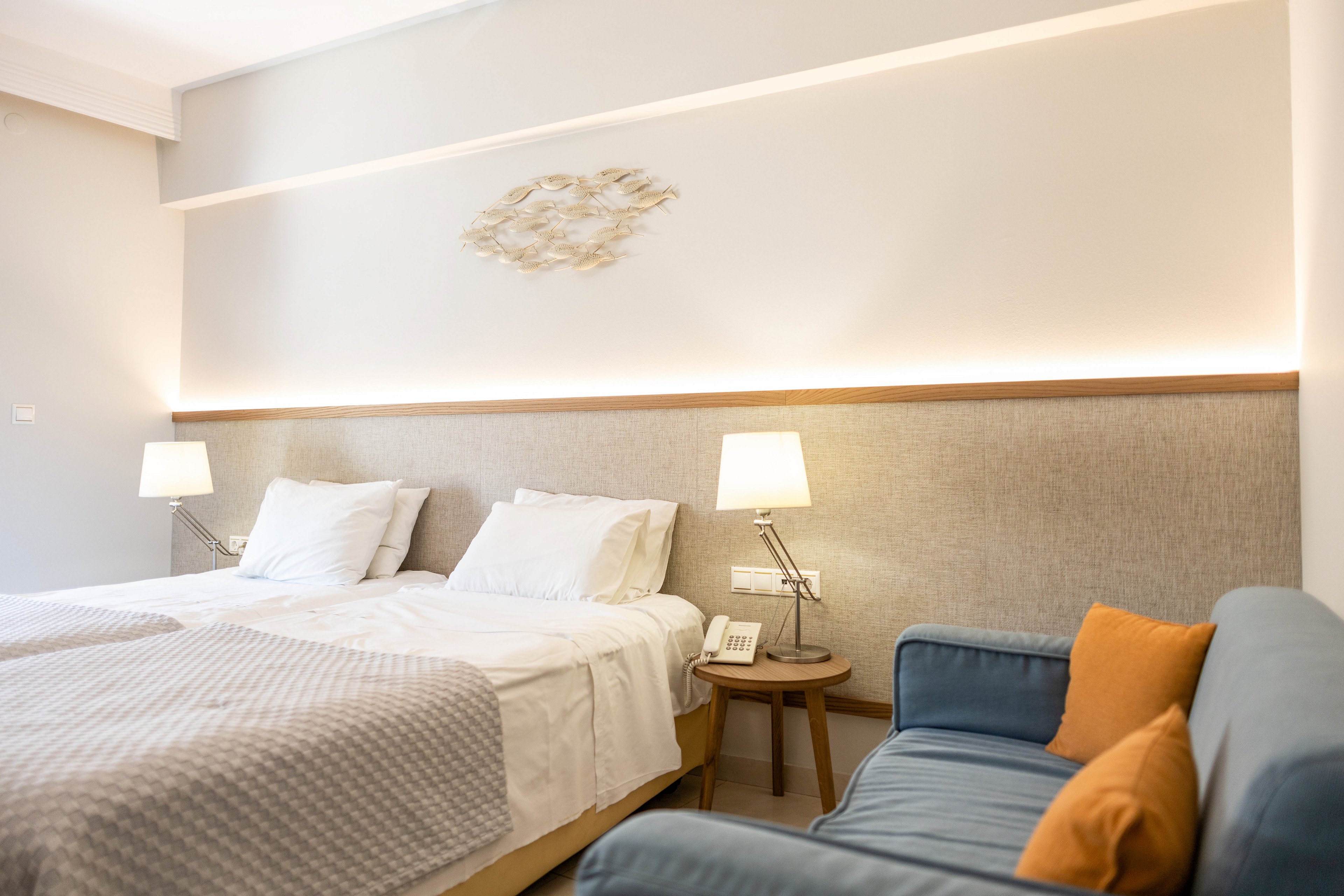 Bright, contemporary hotel room featuring two neatly made twin beds with white linens and textured blankets. A soft upholstered headboard runs the length of the wall, complemented by two bedside tables with matching lamps. Warm accent lighting and minimalist wall decor create a calm, inviting atmosphere. A blue armchair with contrasting orange cushions adds a pop of color to the neutral interior.