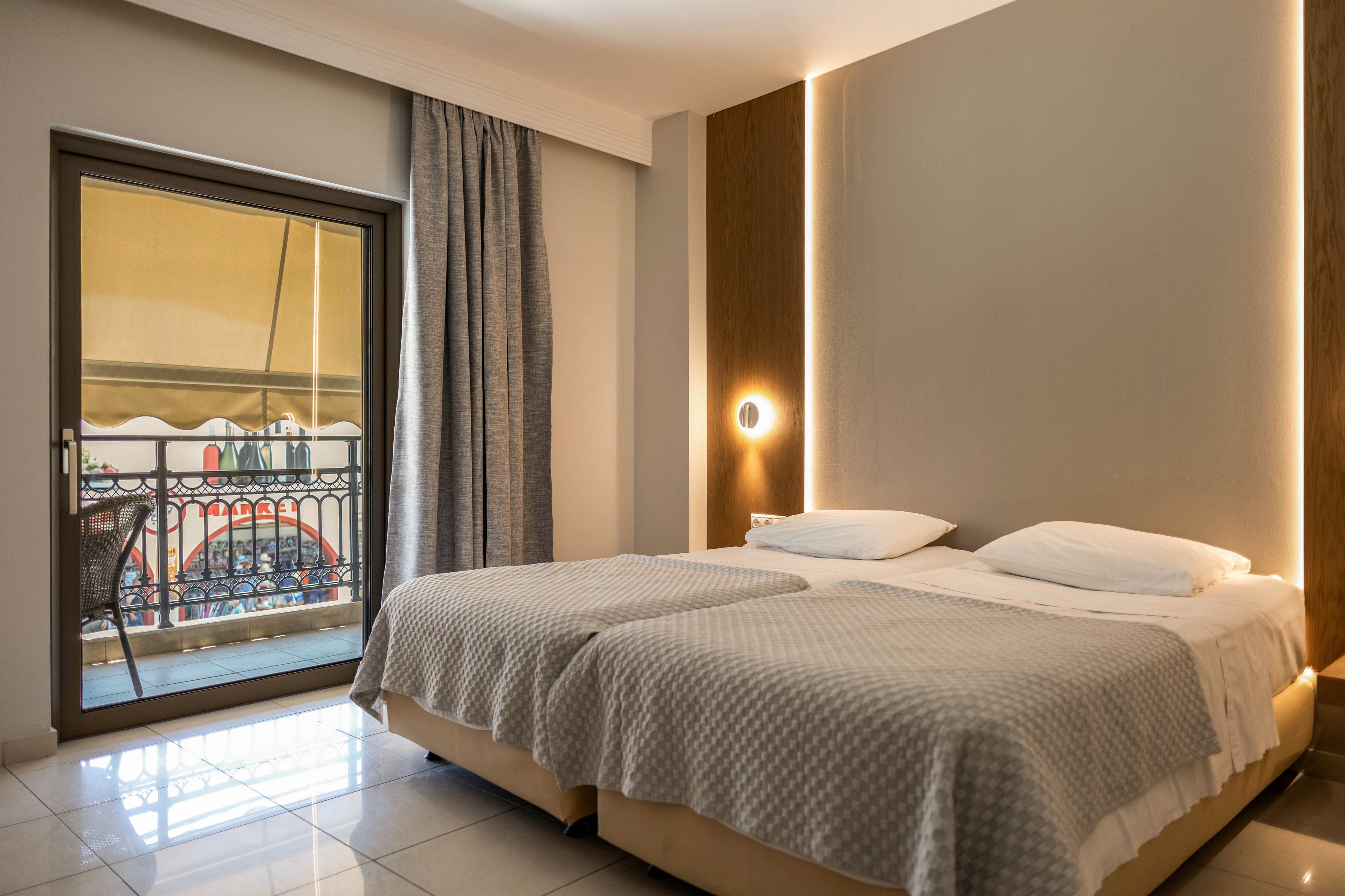 A contemporary hotel room features two neatly made twin beds with light gray textured bedspreads. Warm vertical lighting and a round wall sconce create a cozy atmosphere against neutral walls. A large glass door opens to a balcony with a decorative railing and outdoor seating, allowing natural light to fill the tiled floor interior. Heavy curtains frame the balcony door, adding a touch of elegance and privacy to the space.