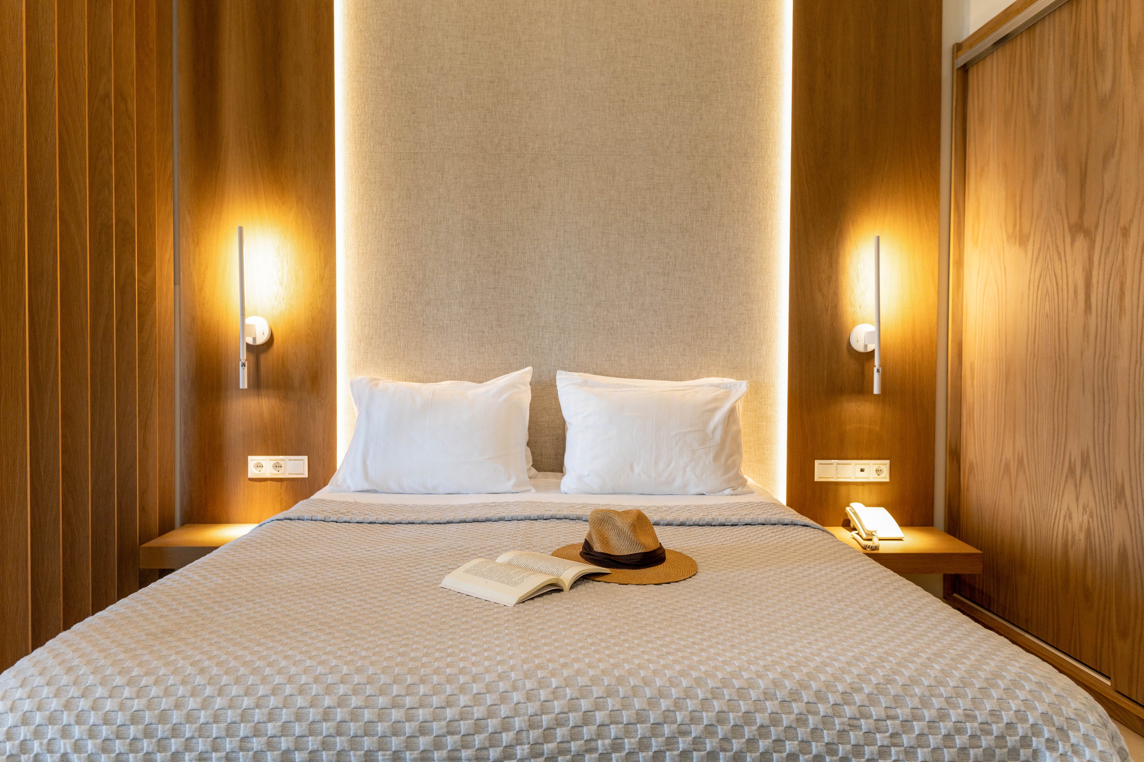 A neatly made double bed with white pillows and a textured light gray bedspread is centered in a modern hotel room. Warm wooden wall panels and vertical bedside lights create a soft, inviting glow around a fabric headboard. An open book and a hat rest on the bed, suggesting relaxation or leisure travel. Minimalist bedside tables with a telephone and wall switches complete the contemporary, cozy setting.