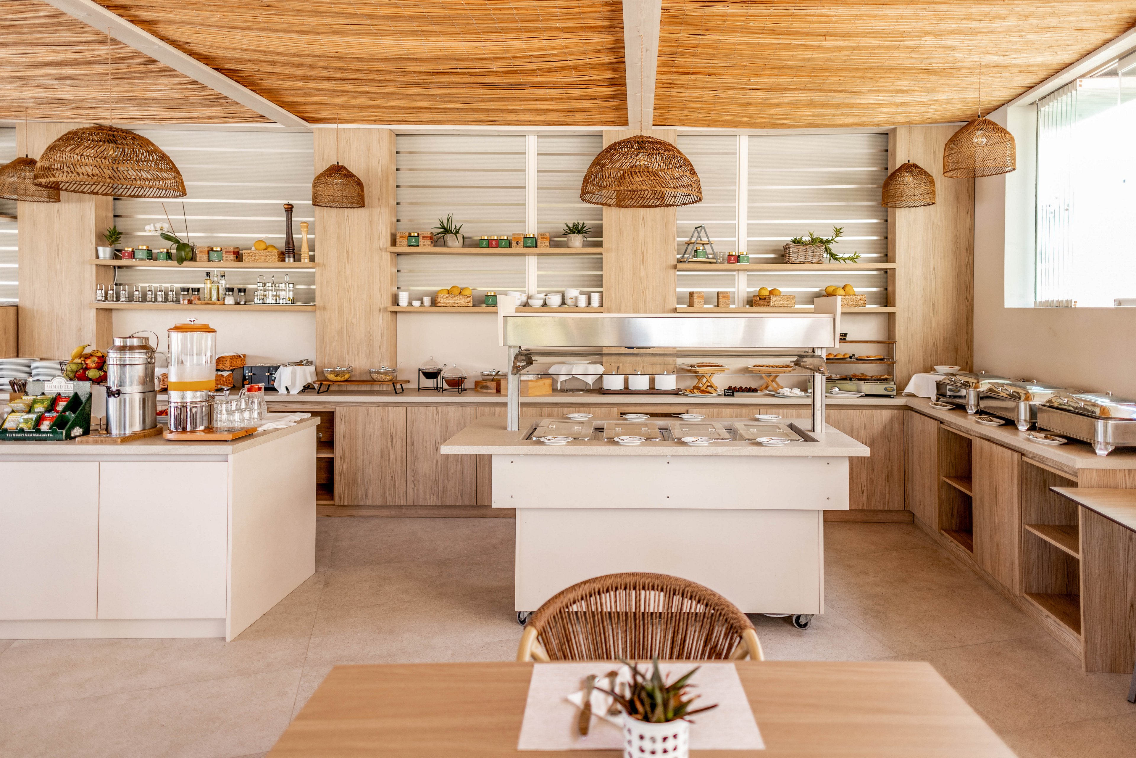 A modern self-service buffet is set up in a bright, airy cafe interior. Wooden shelves and counters display assorted breakfast foods, beverages, and tableware. Woven pendant lights and neutral tones create a warm, natural atmosphere. A foreground table with a small potted plant adds a welcoming touch to the dining area.