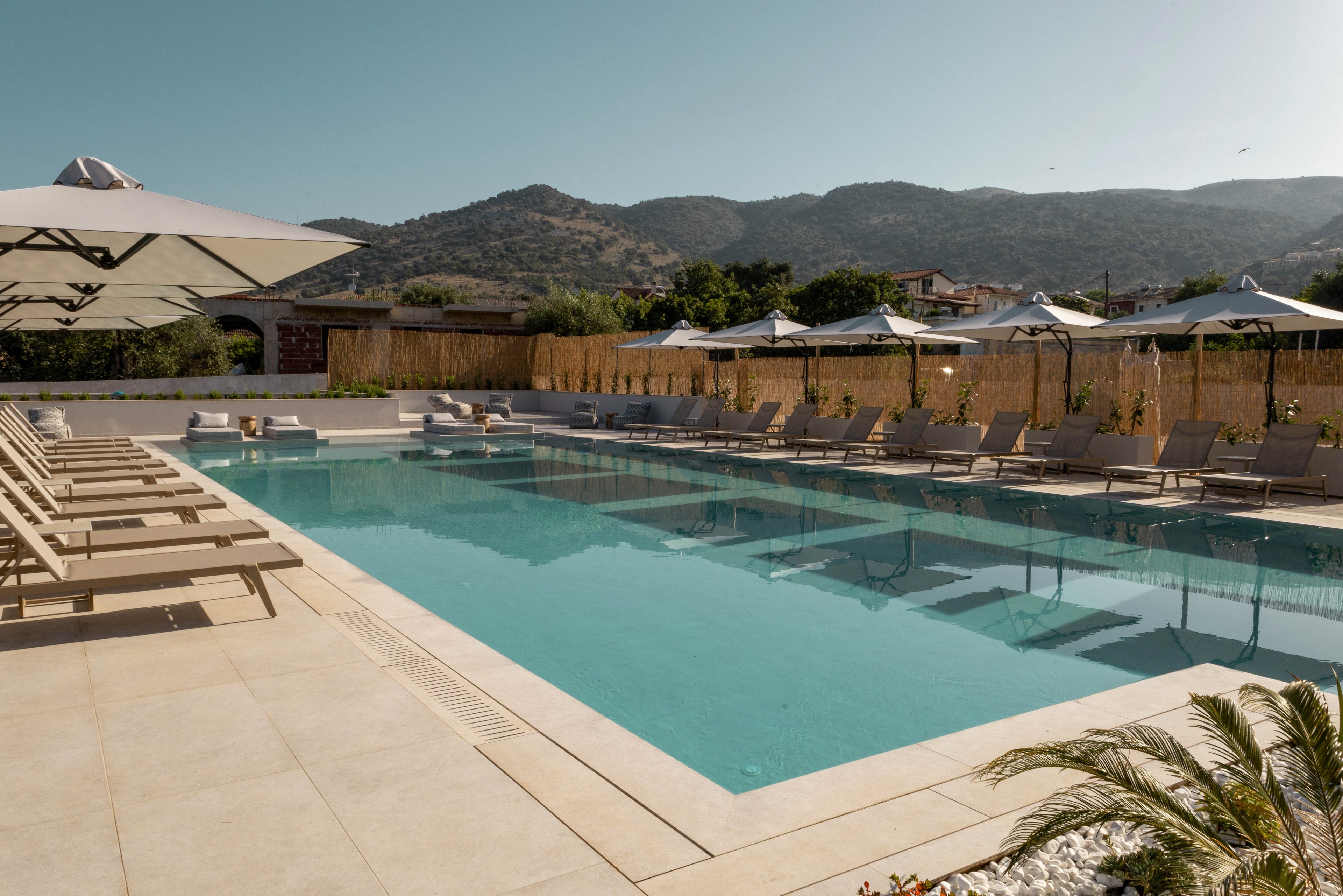 A large rectangular outdoor swimming pool is surrounded by beige lounge chairs and umbrellas in a modern resort setting. The scene is calm and sunlit, with clear blue water reflecting the sky. In the background, low buildings and a wooden fence sit beneath rolling green mountains. The overall atmosphere is relaxed and inviting, ideal for vacation or leisure.