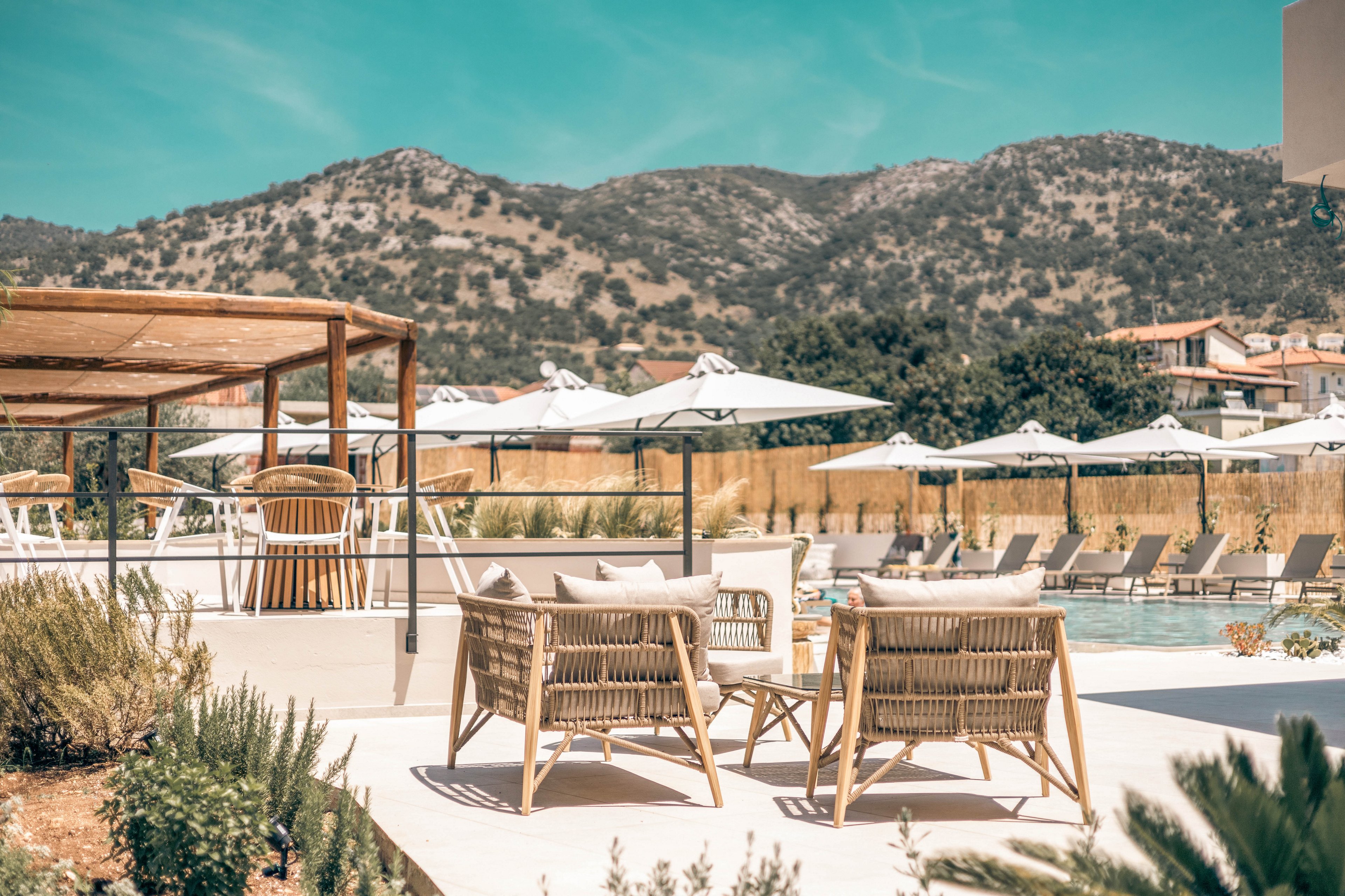 Outdoor lounge chairs and tables sit beside a resort swimming pool under clear turquoise skies. White sun umbrellas line the pool area, offering shade to the seating and sunbeds. A wooden pergola and natural wicker furniture create a relaxed, coastal atmosphere. In the background, tree-covered hills and scattered buildings frame the tranquil vacation setting.