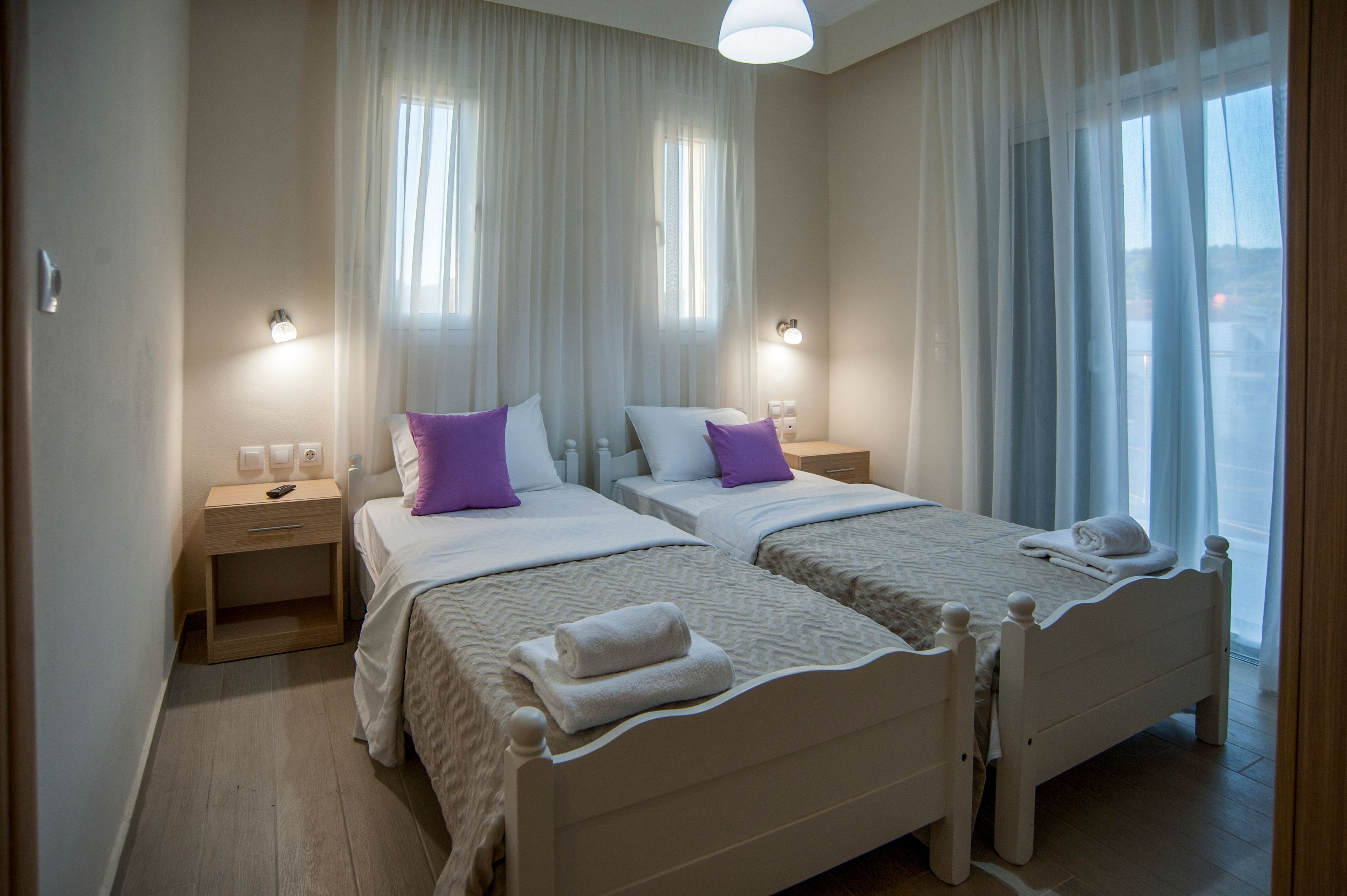 A neatly arranged twin bedroom features two single beds with light-colored frames and matching beige bedspreads. Each bed is topped with folded white towels and a purple accent pillow, adding a subtle pop of color. Sheer curtains cover the windows, allowing diffused daylight to soften the room, while wall-mounted lamps and a ceiling light provide additional illumination. Wooden nightstands with electrical outlets and switches emphasize the room’s functional, modern design.