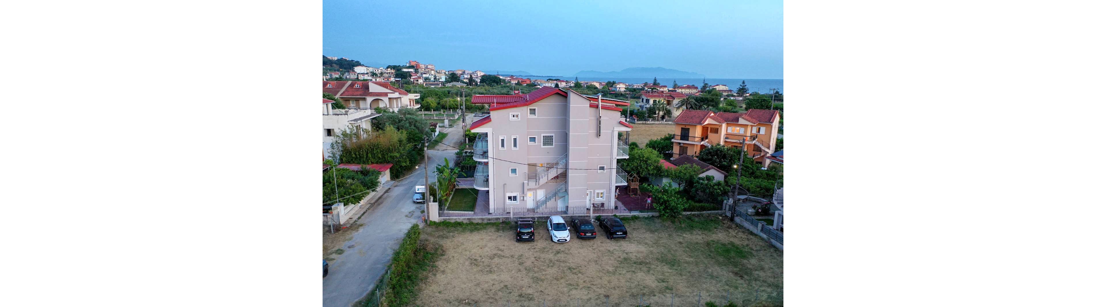 An aerial view shows a multi-story apartment building with a red roof in a suburban neighborhood. Several cars are parked in front of the building along a small dirt lot. Surrounding the property are other houses, greenery, and narrow streets leading toward the distant coastline. The sky is clear and blue, suggesting a calm evening atmosphere.