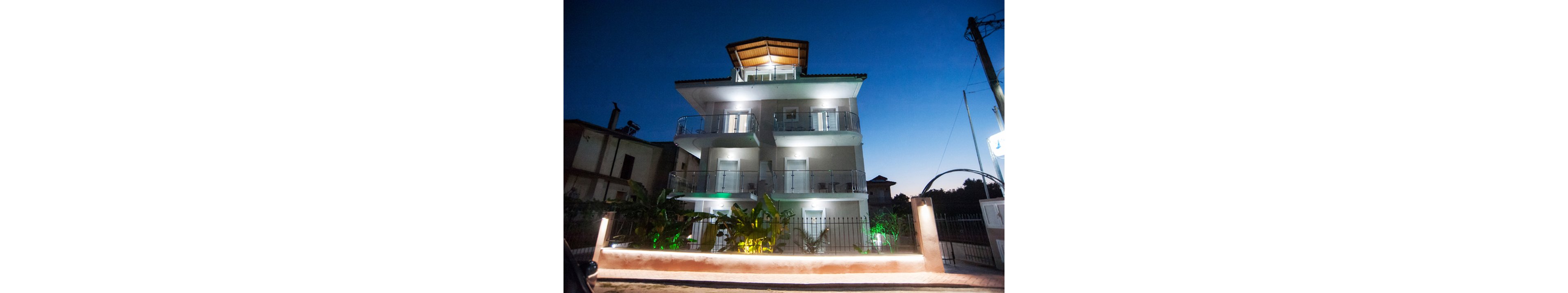 A modern three-story residential building is brightly illuminated against a deep blue evening sky. The structure features multiple balconies with glass railings and a prominent wooden roof overhang. Decorative exterior lighting highlights the white facade and landscaped plants along the front fence. A low wall and metal gate frame the property, with neighboring buildings faintly visible in the background.