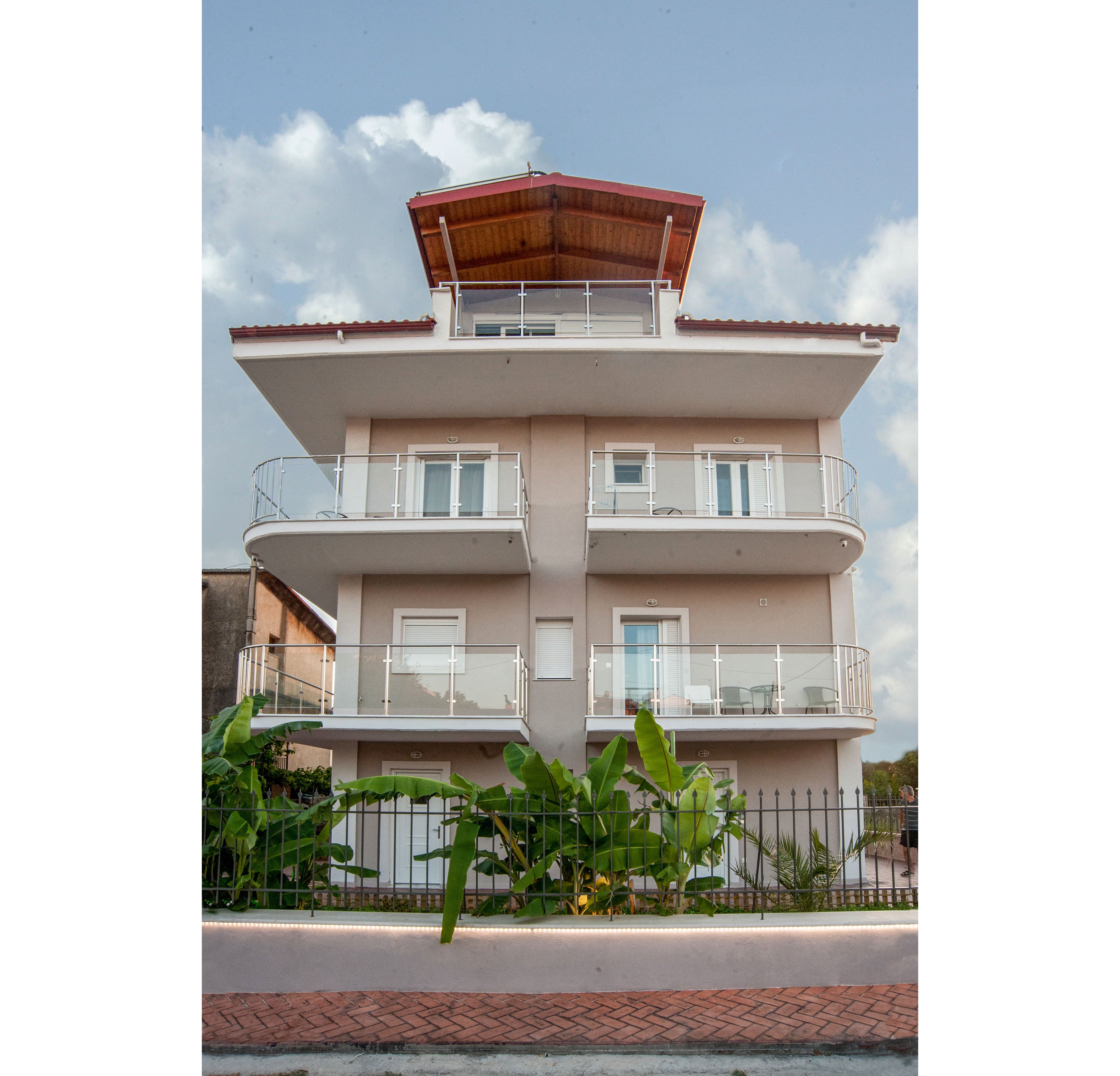 A modern three-story residential building with multiple balconies stands behind a low fence and sidewalk. The structure features light-colored walls, white railings, and large windows, topped with a wooden-roofed penthouse level. Green plants and small trees grow in front of the building, adding contrast to the neutral facade. The scene is captured in daylight under a partly cloudy sky, emphasizing the clean architectural lines and contemporary design.