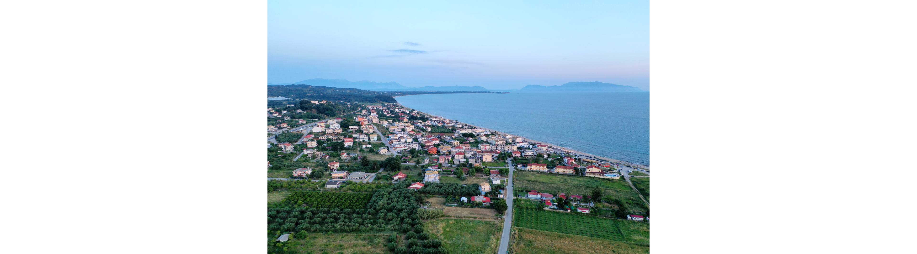 An aerial view captures a small coastal town stretching along a calm shoreline. Residential buildings and houses are arranged in neat blocks near the beach, with agricultural fields and orchards in the foreground. The sea appears tranquil, and distant hills and headlands frame the horizon under a soft evening sky. The overall scene conveys a peaceful seaside community surrounded by greenery.