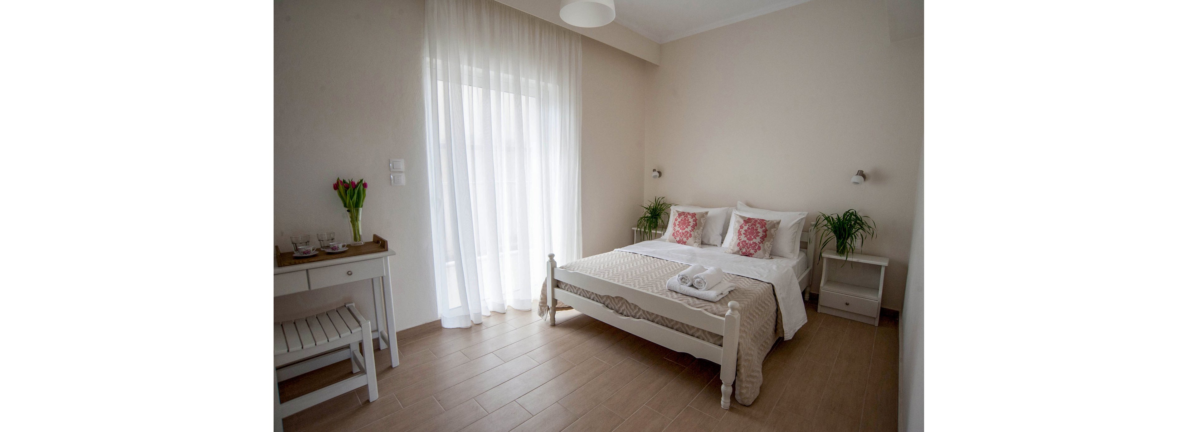 A bright, minimalist bedroom features a double bed with white linens and decorative pink pillows. Soft natural light filters through sheer floor-to-ceiling curtains covering a large window or balcony door. A small desk with a chair and vase of red tulips sits to the left, while potted plants add greenery on either side of the bed. Neutral beige walls and light wood flooring create a calm, airy atmosphere suitable for rest or a guest room.