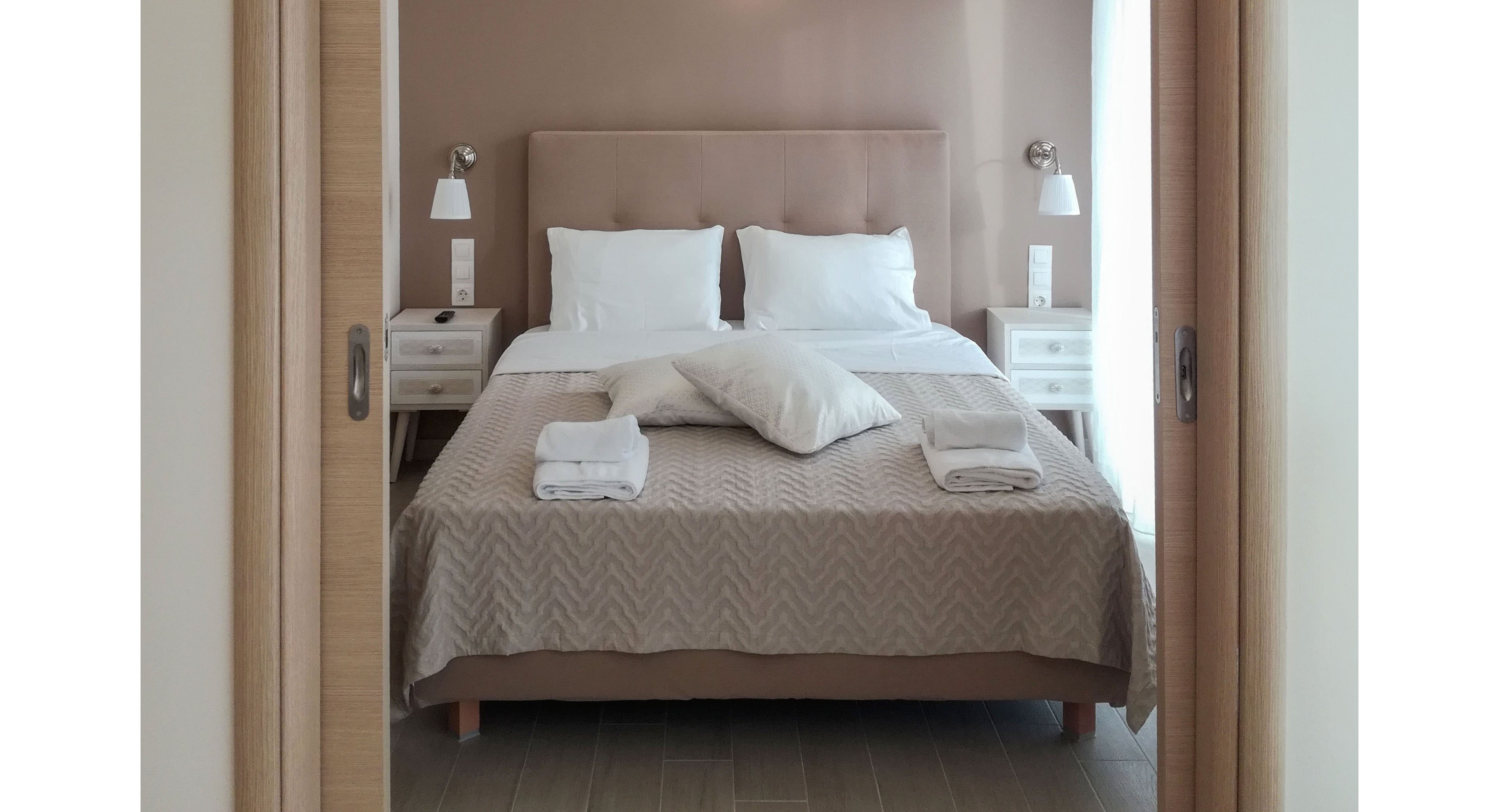 A neatly made double bed is centered in a minimalist bedroom, viewed through partially open sliding doors. The bed features white pillows, folded towels, and a neutral-toned bedspread against a soft beige accent wall. Matching bedside tables with lamps and switches frame the bed, creating a symmetrical composition. Natural light filters in from the side, giving the room a calm and inviting atmosphere.