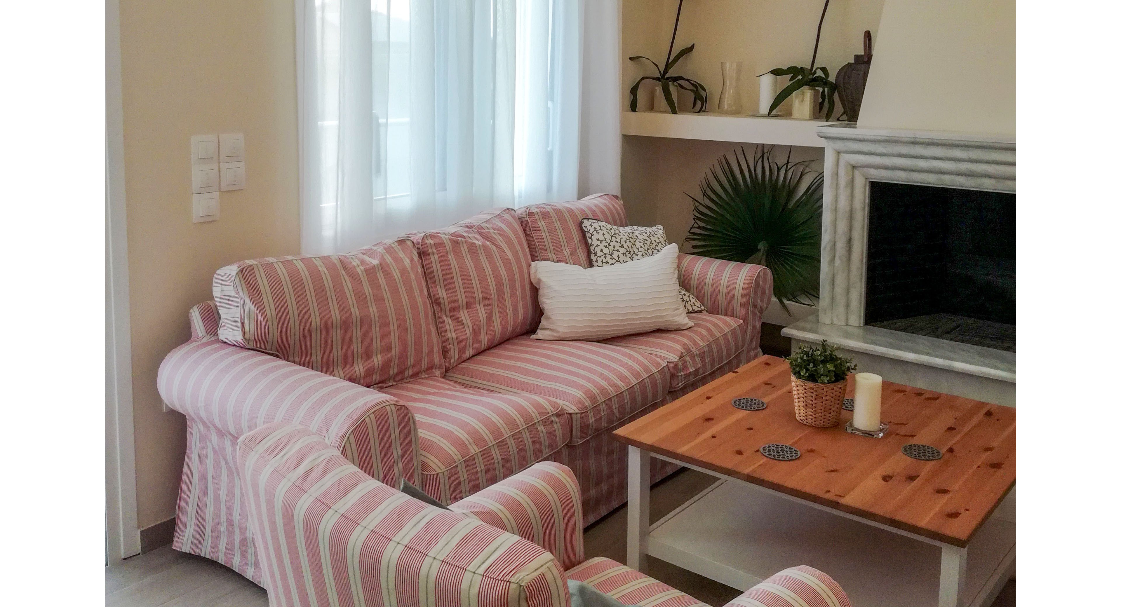A cozy living room features a pink and white striped sofa and matching armchair arranged around a wooden coffee table. Soft natural light filters through sheer curtains covering a large window. A marble fireplace with decorative plants and greenery adds a focal point to the space. Neutral walls and simple decor create a calm, inviting atmosphere.