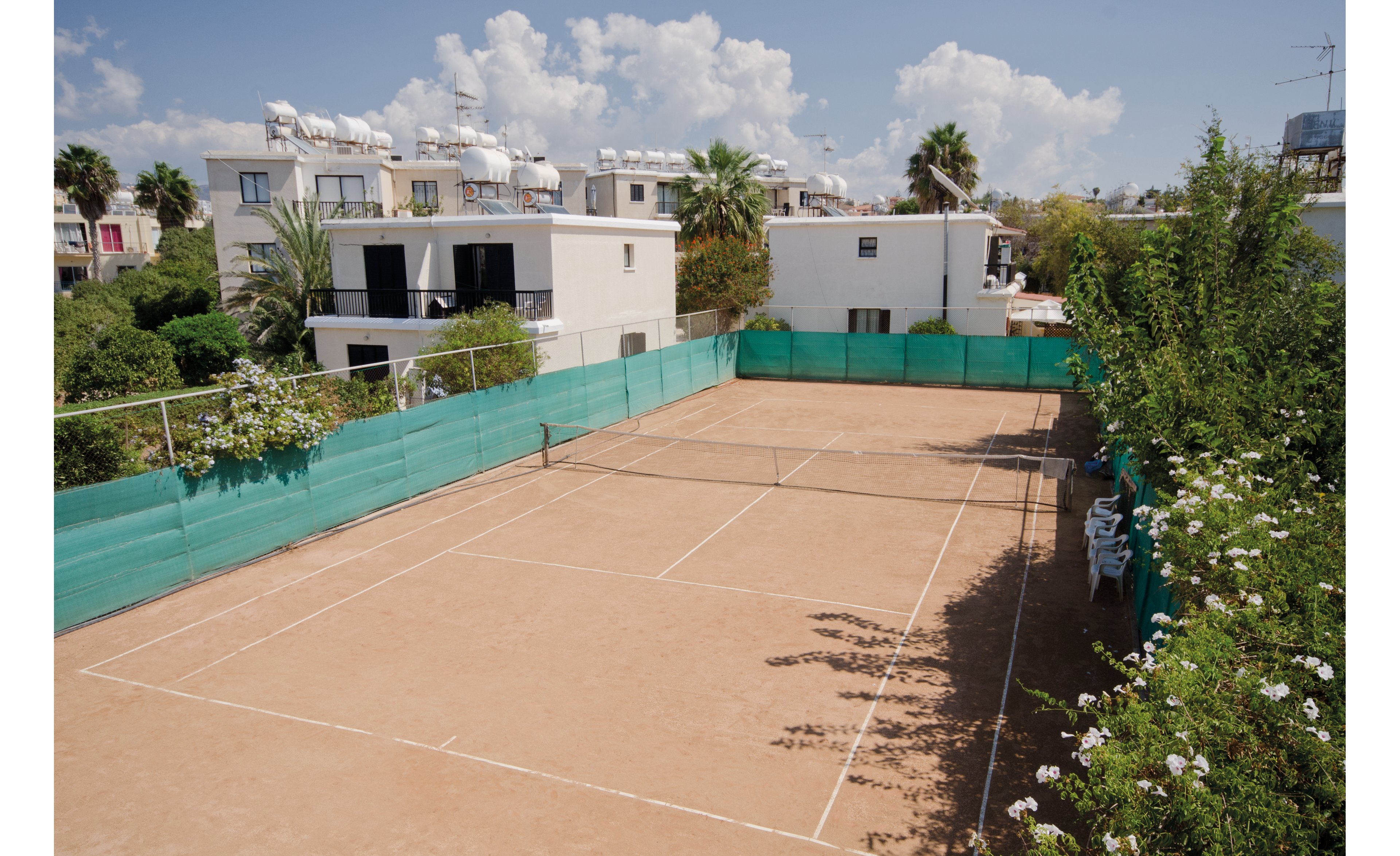 An empty clay tennis court surrounded by green fencing is situated in a sunny residential neighborhood. The setting features modern white buildings with rooftop water tanks and lush greenery. The clear blue sky and scattered clouds add to the serene atmosphere.