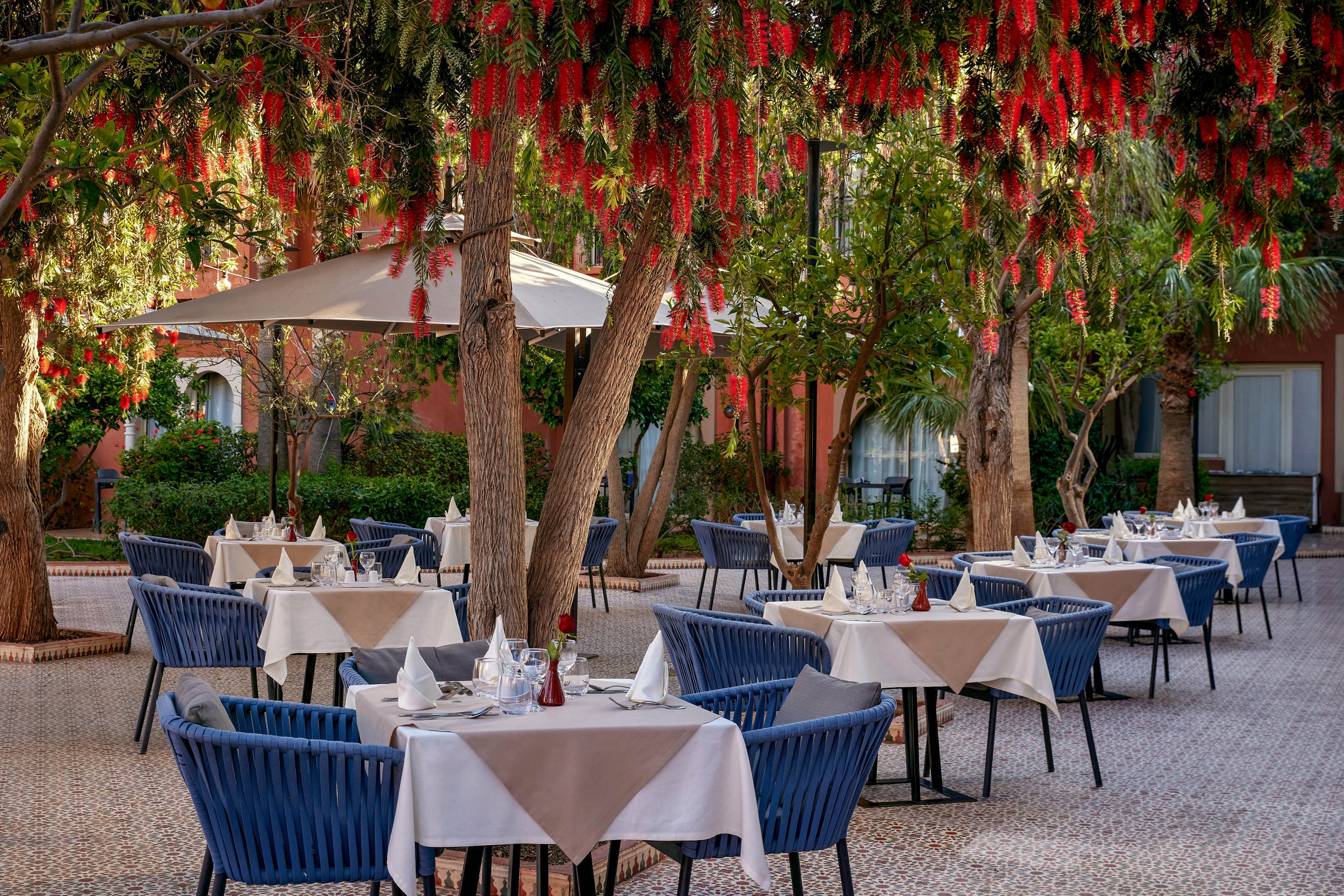 An inviting outdoor restaurant terrace features neatly arranged tables covered with white and beige tablecloths. Blue woven chairs surround each table, which is set with glassware, folded napkins, and condiments. Large umbrellas provide shade beneath lush trees with hanging red flowers, creating a cozy garden atmosphere. The tiled floor and warm building facade in the background add to the relaxed, upscale dining setting.