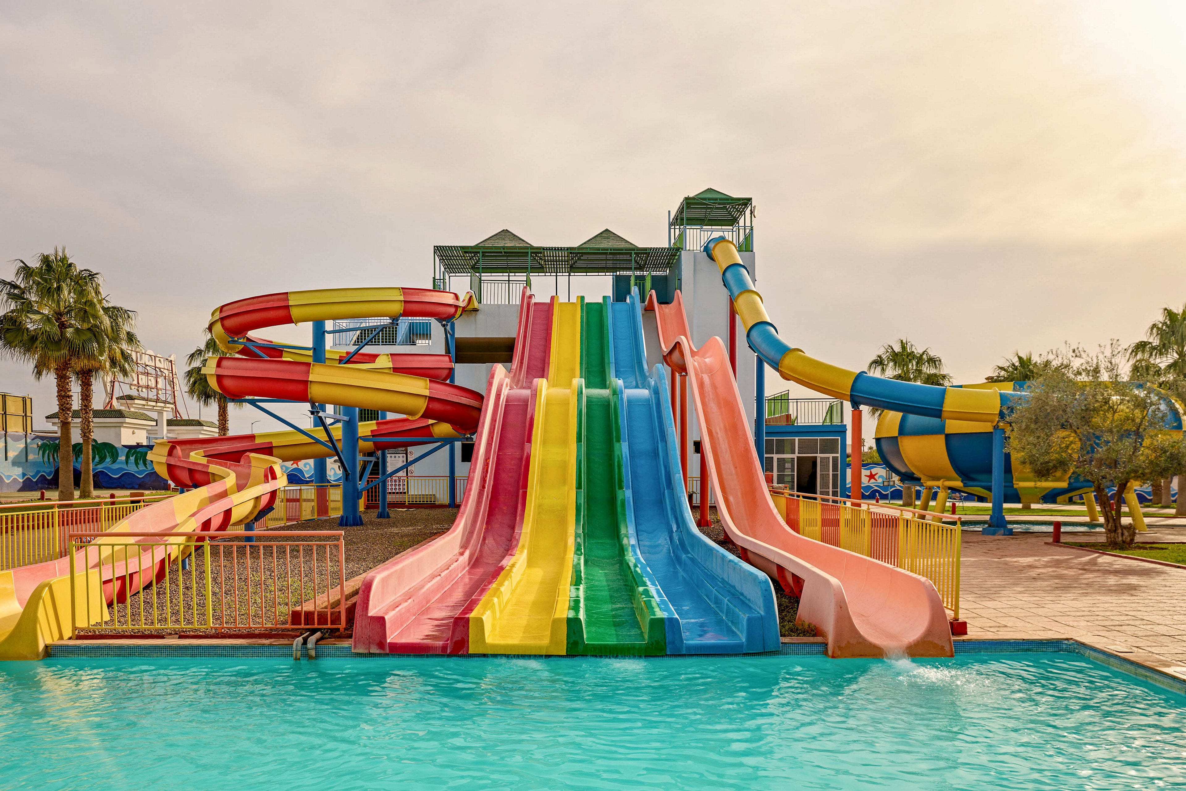 A group of bright, multicolored water slides descends into a clear blue swimming pool at an outdoor water park. The scene includes straight and spiral slides in red, yellow, green, blue, and orange, framed by safety railings. Palm trees and additional park structures are visible in the background under a warm, slightly cloudy sky. The overall atmosphere suggests a fun, family-friendly recreational setting.