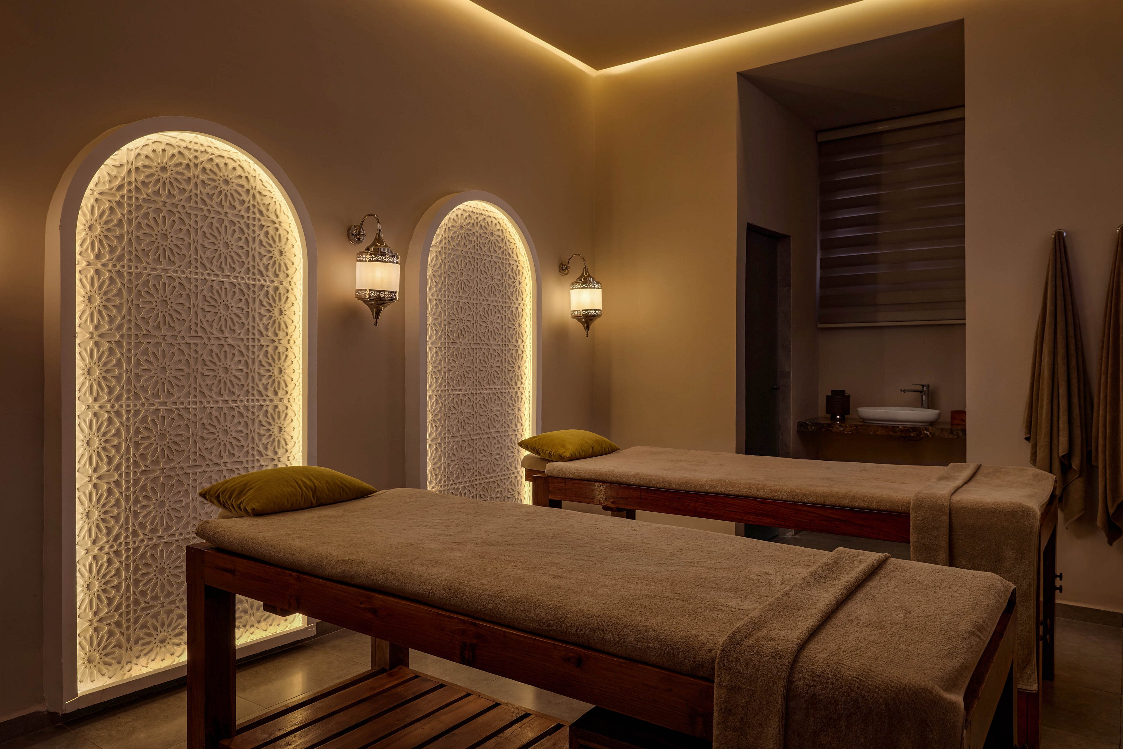 A tranquil spa massage room features two wooden massage tables covered with soft beige linens and green pillows. Warm ambient lighting glows from arched wall niches with decorative patterns, creating a cozy and intimate atmosphere. Wall-mounted lanterns and neatly hung towels enhance the sense of relaxation and comfort. The overall scene suggests a serene space designed for wellness treatments and peaceful retreat.