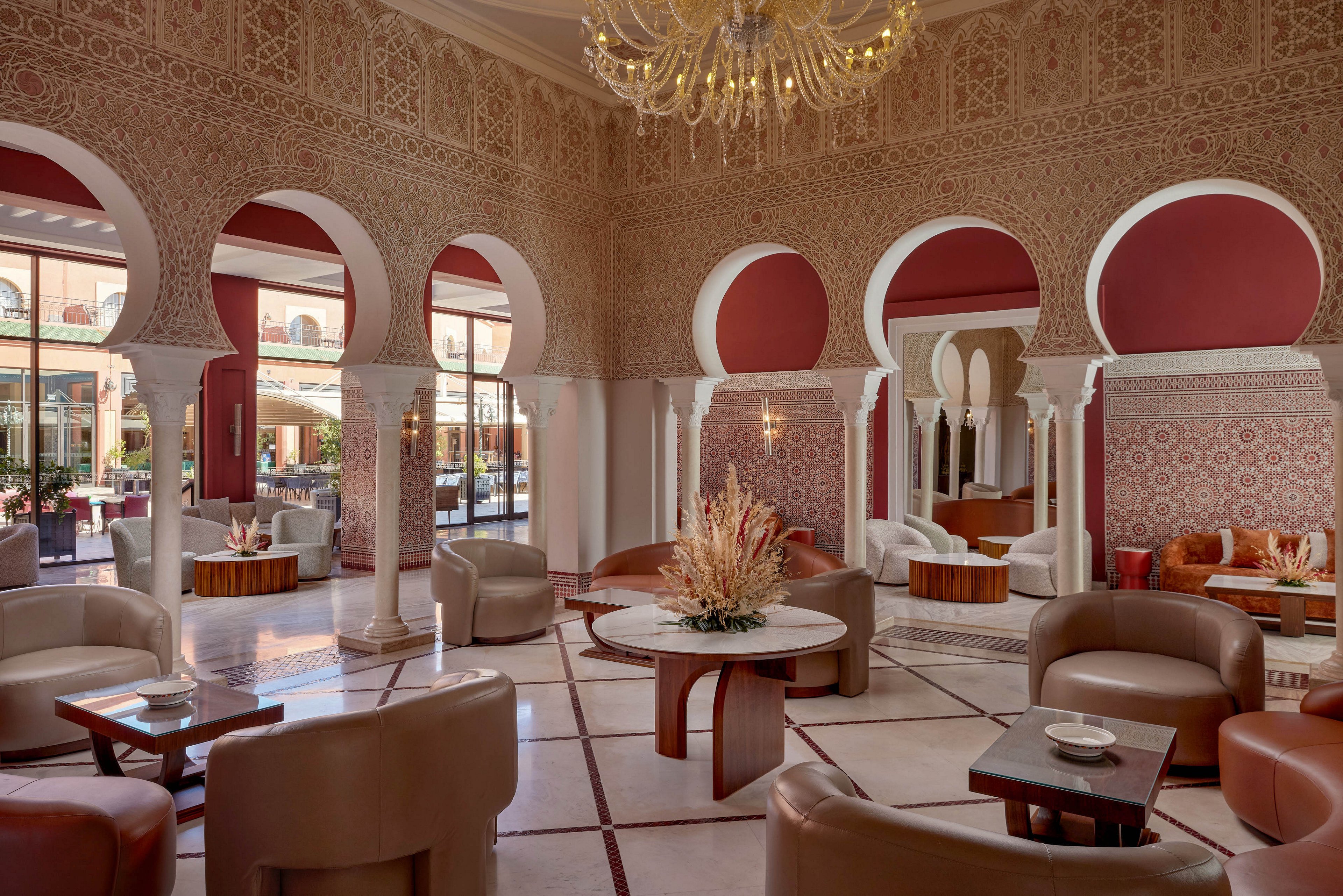 Luxurious lounge seating is arranged beneath grand arches in a spacious hotel interior. Ornate Moroccan-style patterns decorate the walls and columns, complemented by warm red and cream tones. Contemporary armchairs and round tables create an inviting social area, with dried floral arrangements adding texture. Large windows and glass doors bring in natural light and views of the outdoor courtyard.