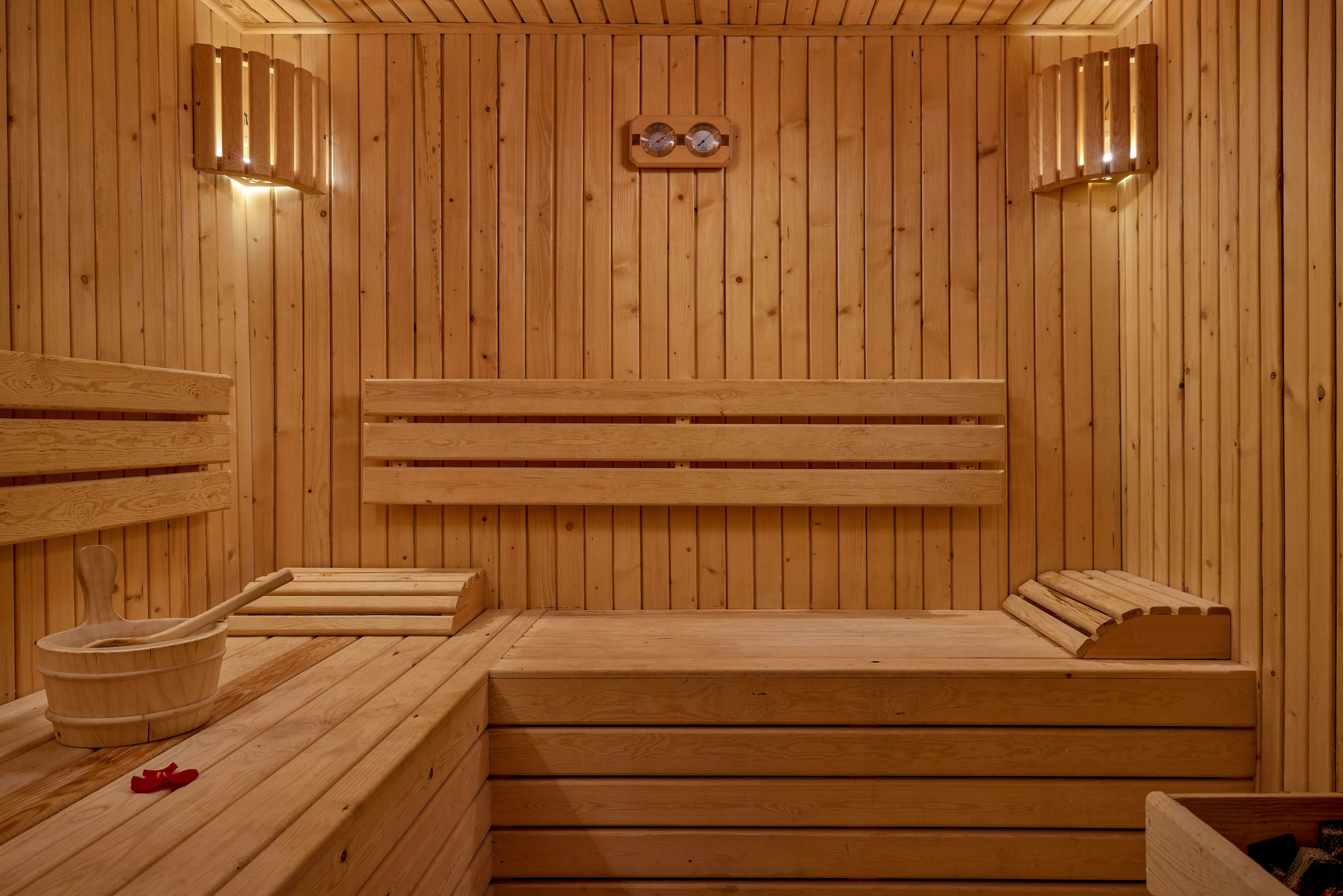 A warm, inviting wooden sauna interior is shown with tiered benches along the walls. Soft lighting fixtures illuminate the vertical wood paneling, creating a cozy atmosphere. A wooden bucket and ladle sit on the lower bench, emphasizing the traditional sauna experience. The space is clean, minimal, and ready for relaxation.