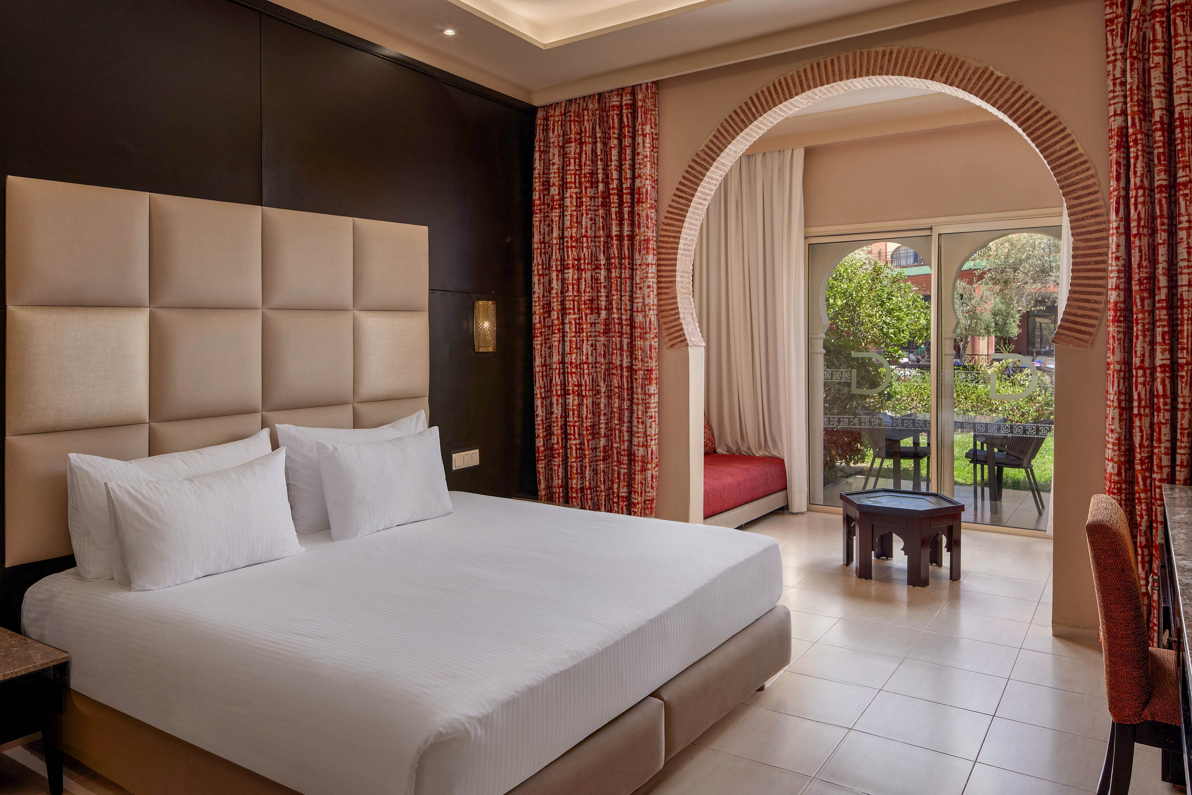 A contemporary hotel room features a large bed with padded headboard and crisp white linens. The space includes patterned red curtains, a cozy window seat, and neutral tiled flooring. An arched doorway opens onto a terrace with outdoor chairs and a small table, overlooking a lush garden. Warm lighting and soft colors create a comfortable, inviting atmosphere.
