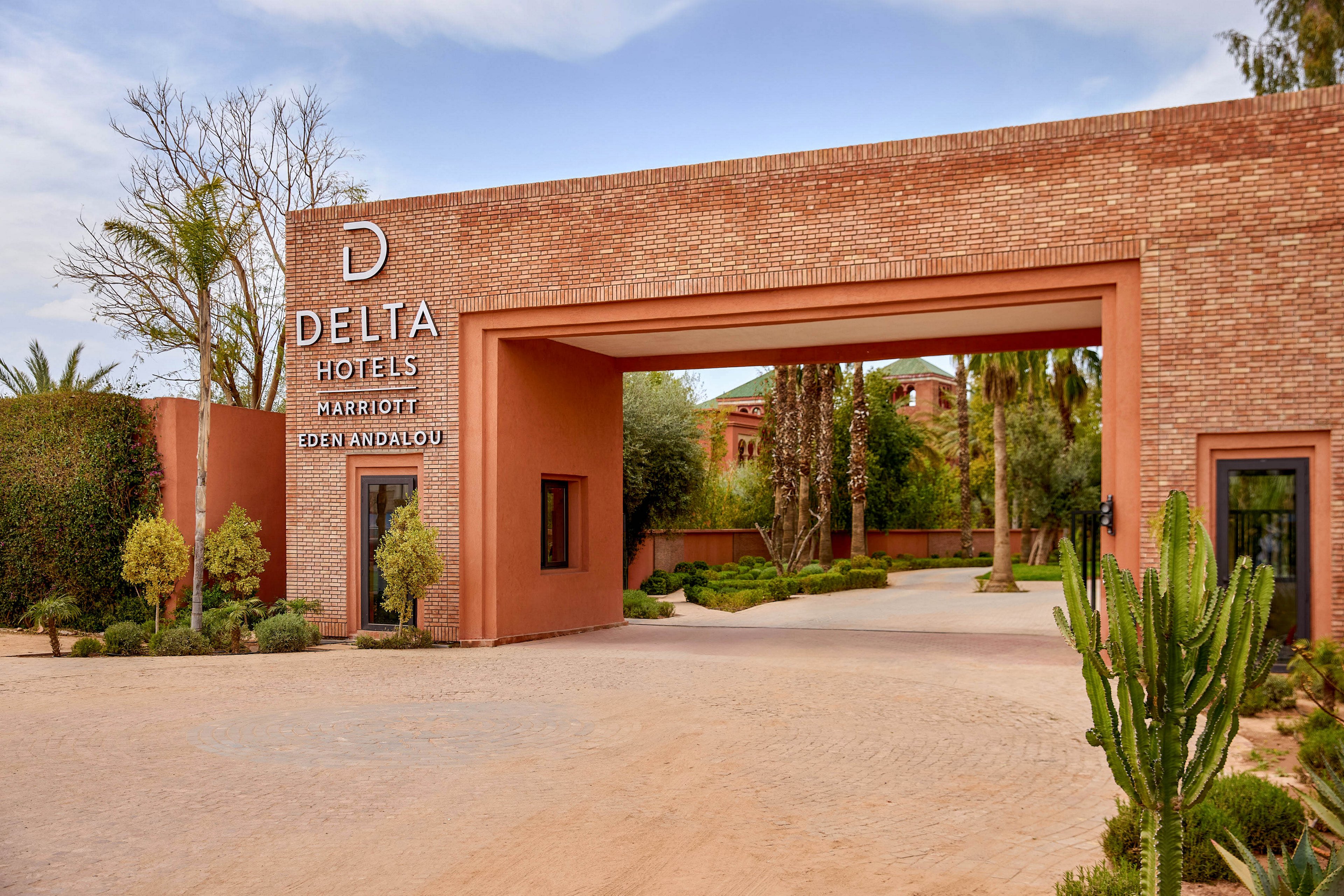 A brick archway marks the entrance to Delta Hotels Marriott Eden Andalou. The scene shows a landscaped driveway with desert plants and manicured shrubs leading toward the hotel buildings. Warm terracotta tones dominate the architecture, complemented by greenery and a clear sky. The setting suggests a tranquil resort environment in a sunny, dry climate.