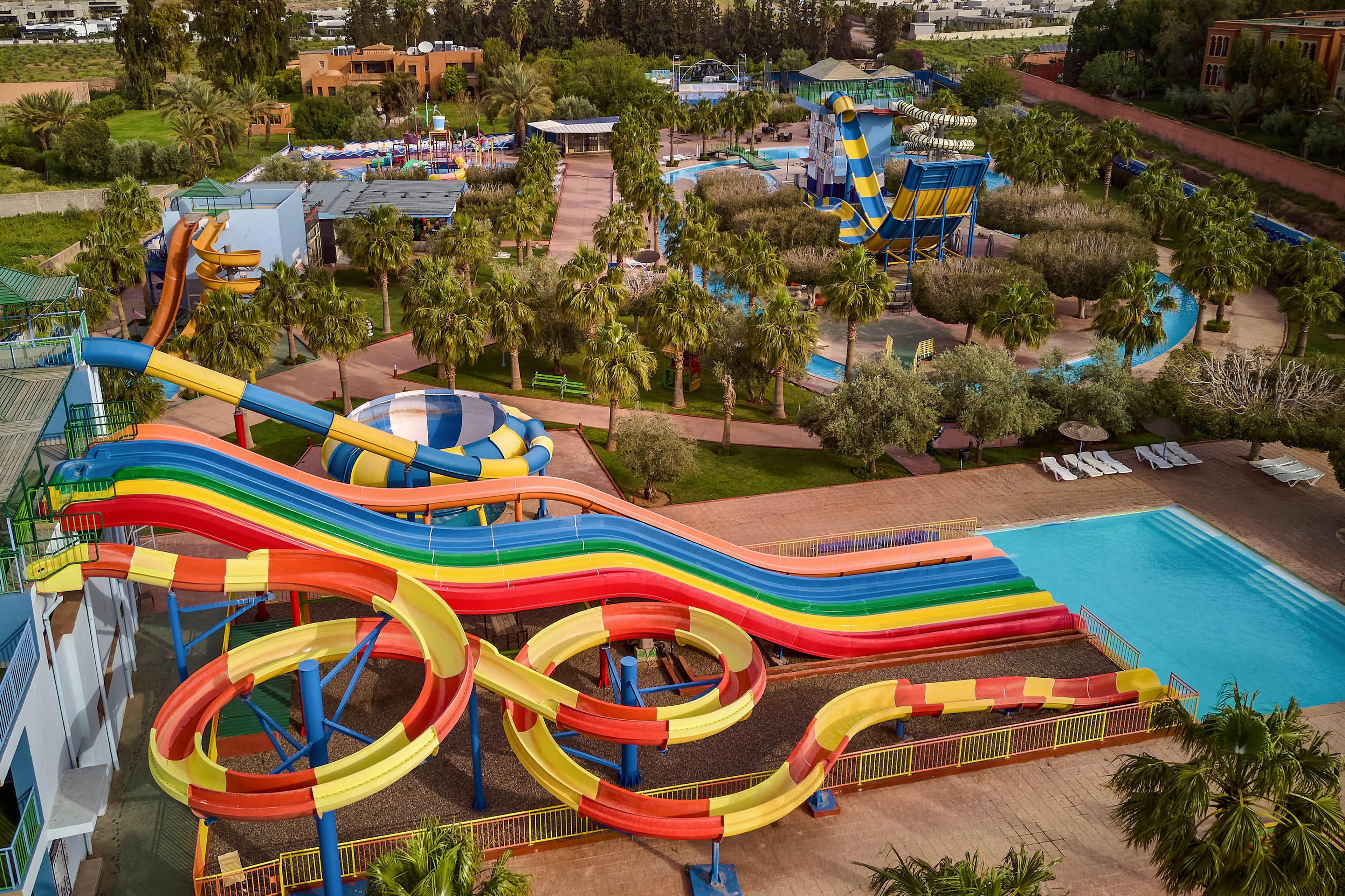 A large outdoor water park features multiple bright, multicolored slides winding above a clear blue swimming pool. The scene is surrounded by palm trees, landscaped greenery, and pathways leading to other attractions. In the background, additional water slides, play areas, and seating zones are visible, suggesting a family-friendly recreational setting. The overall atmosphere is vibrant, sunny, and inviting for leisure and fun.